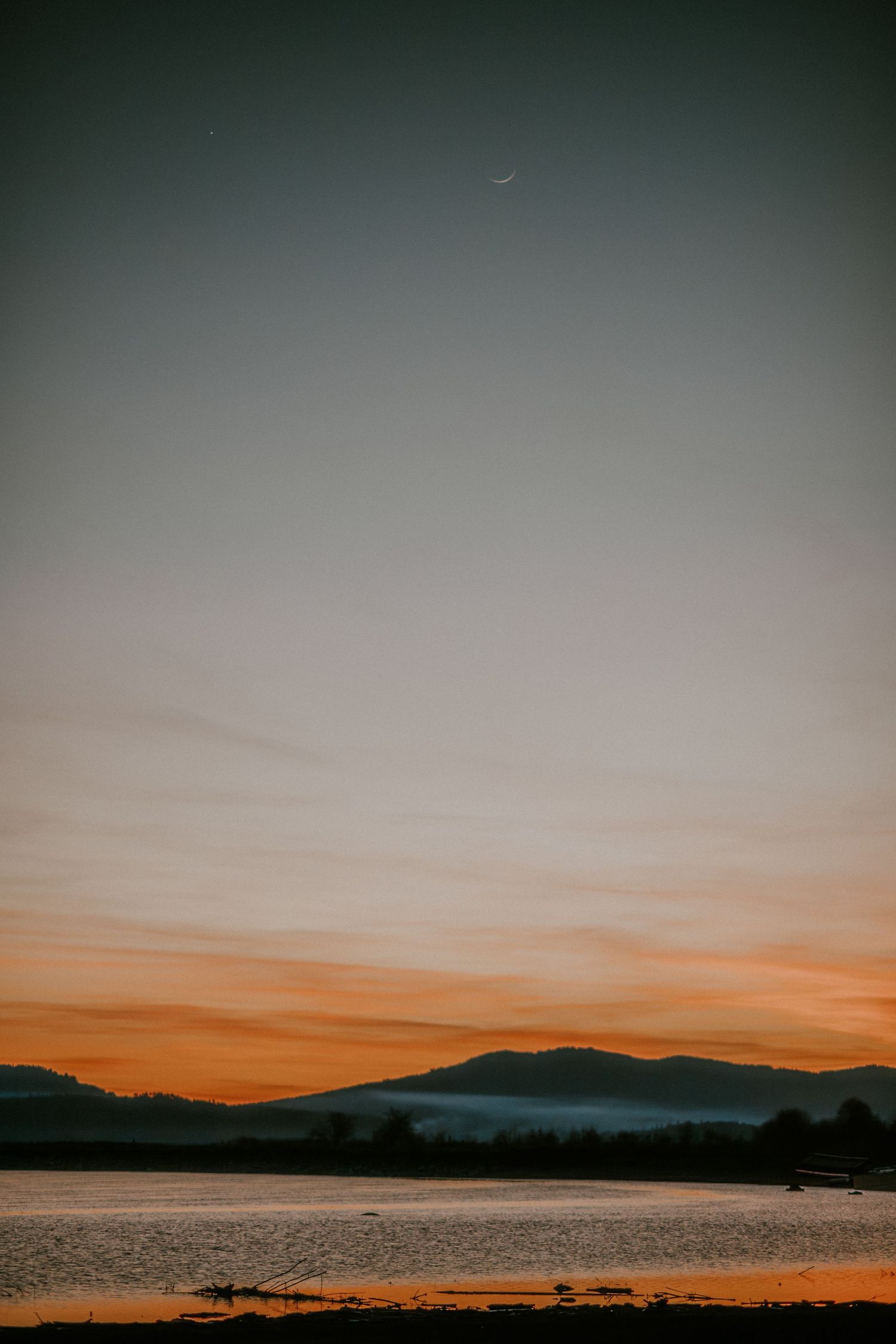 A stunning sunset over a calm lake with silhouetted mountains and a crescent moon in the twilight sky.