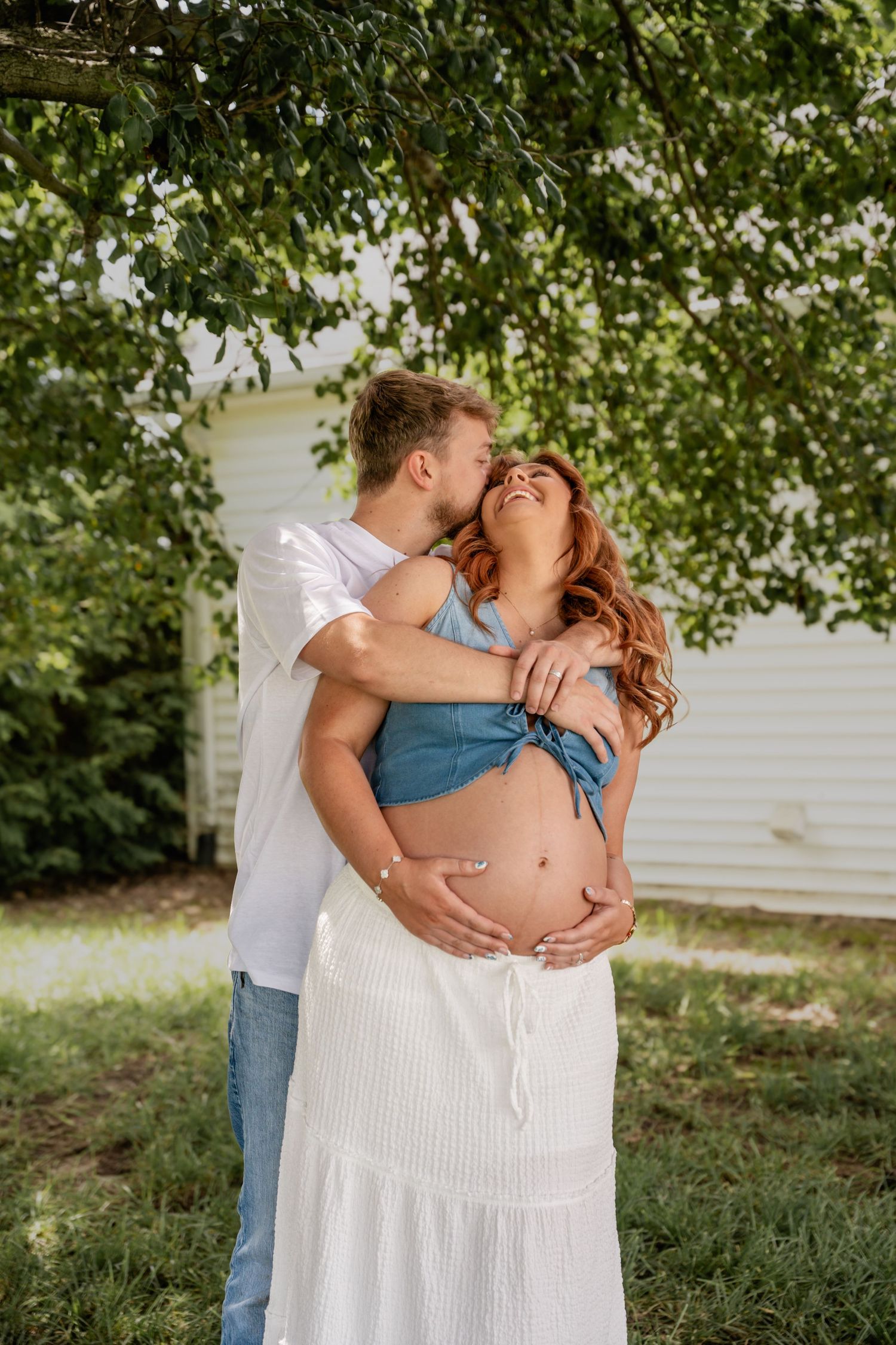 Couple embracing outdoors under green tree, man kissing pregnant woman's cheek while holding her belly.