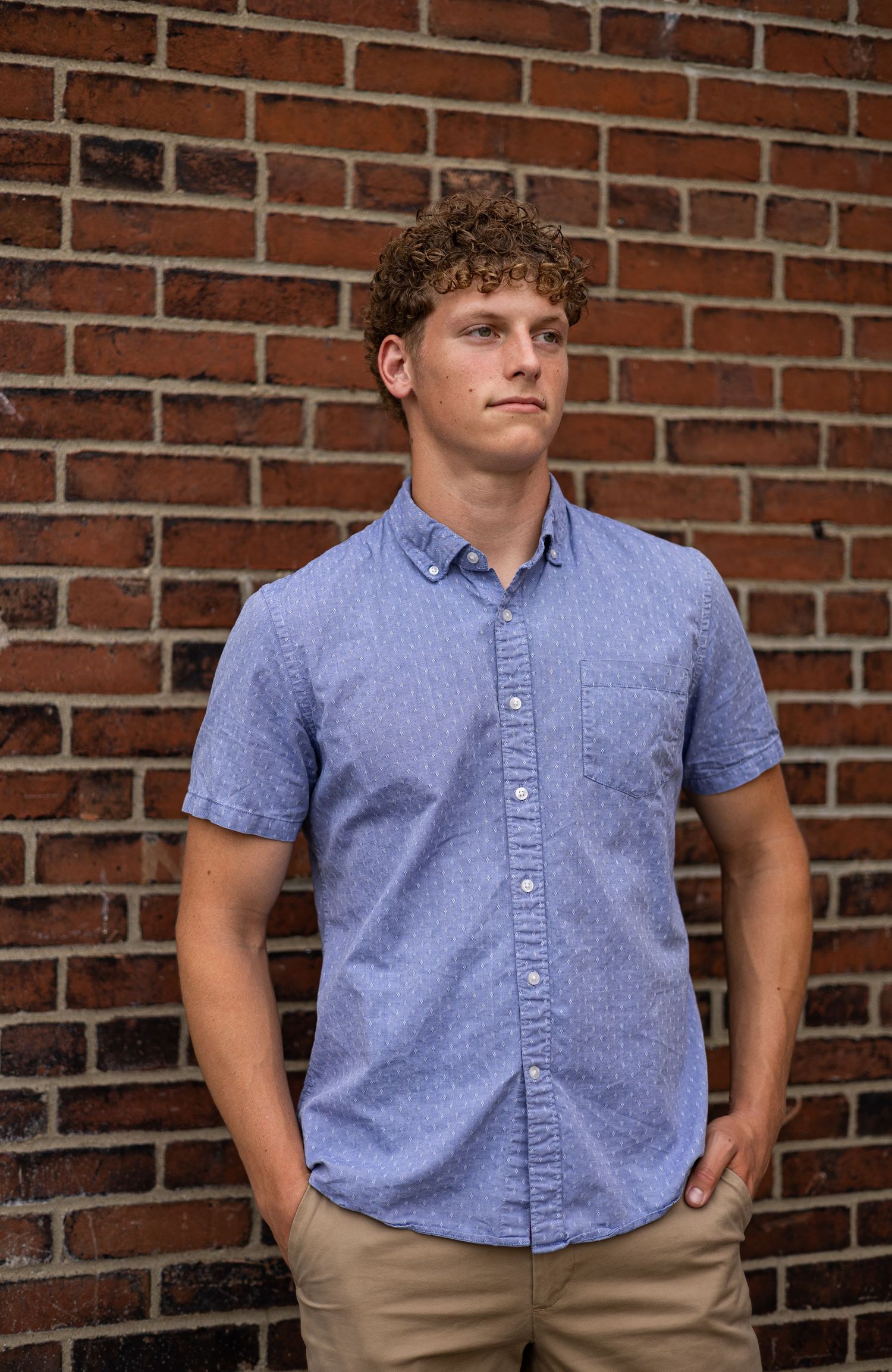 Young man with curly hair posing confidently against a red brick wall in a blue shirt.