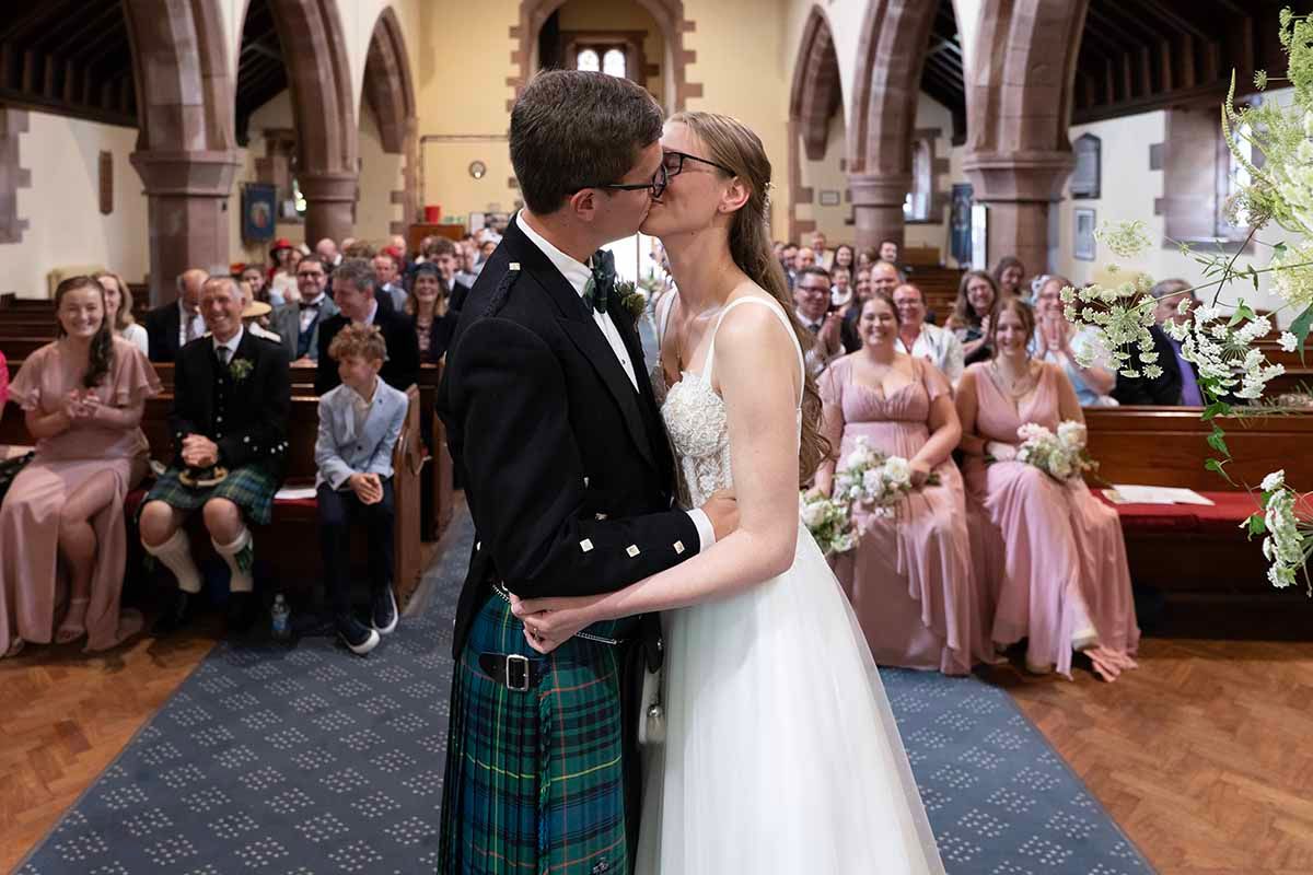 Bride and groom share first kiss at church wedding altar, groom wearing Scottish tartan kilt, guests smiling in background.