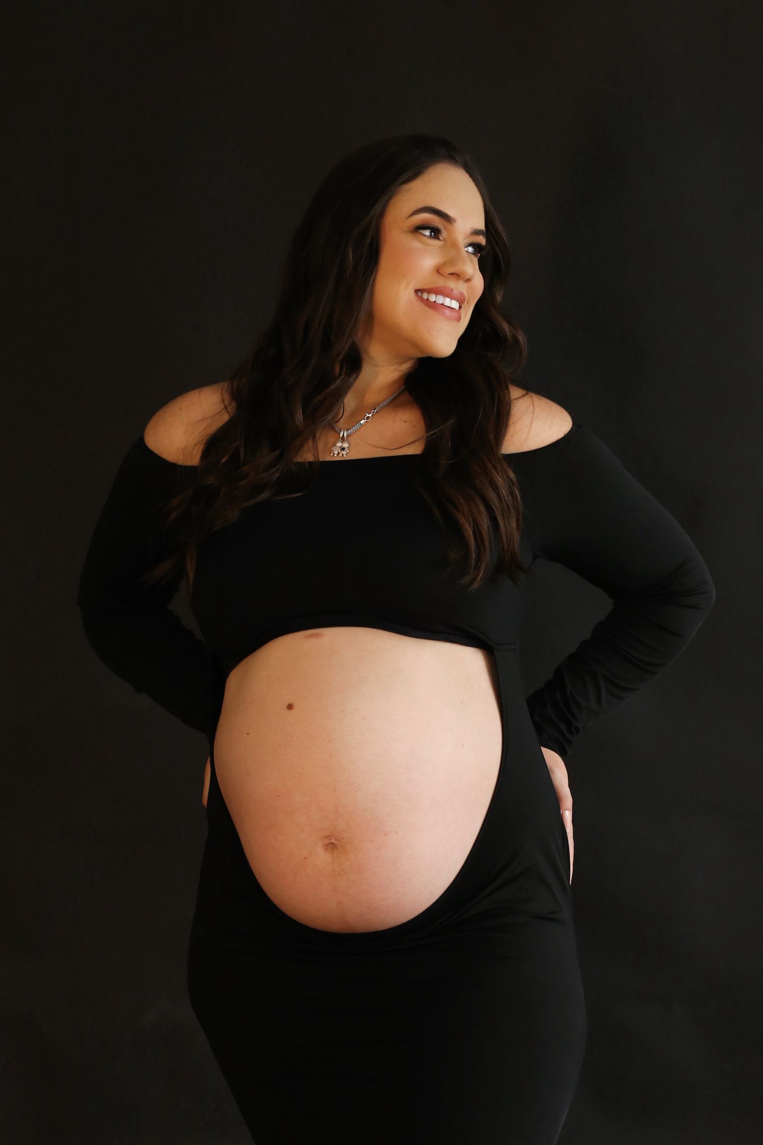 Smiling pregnant woman in black off-shoulder outfit showcasing baby bump against dark background.