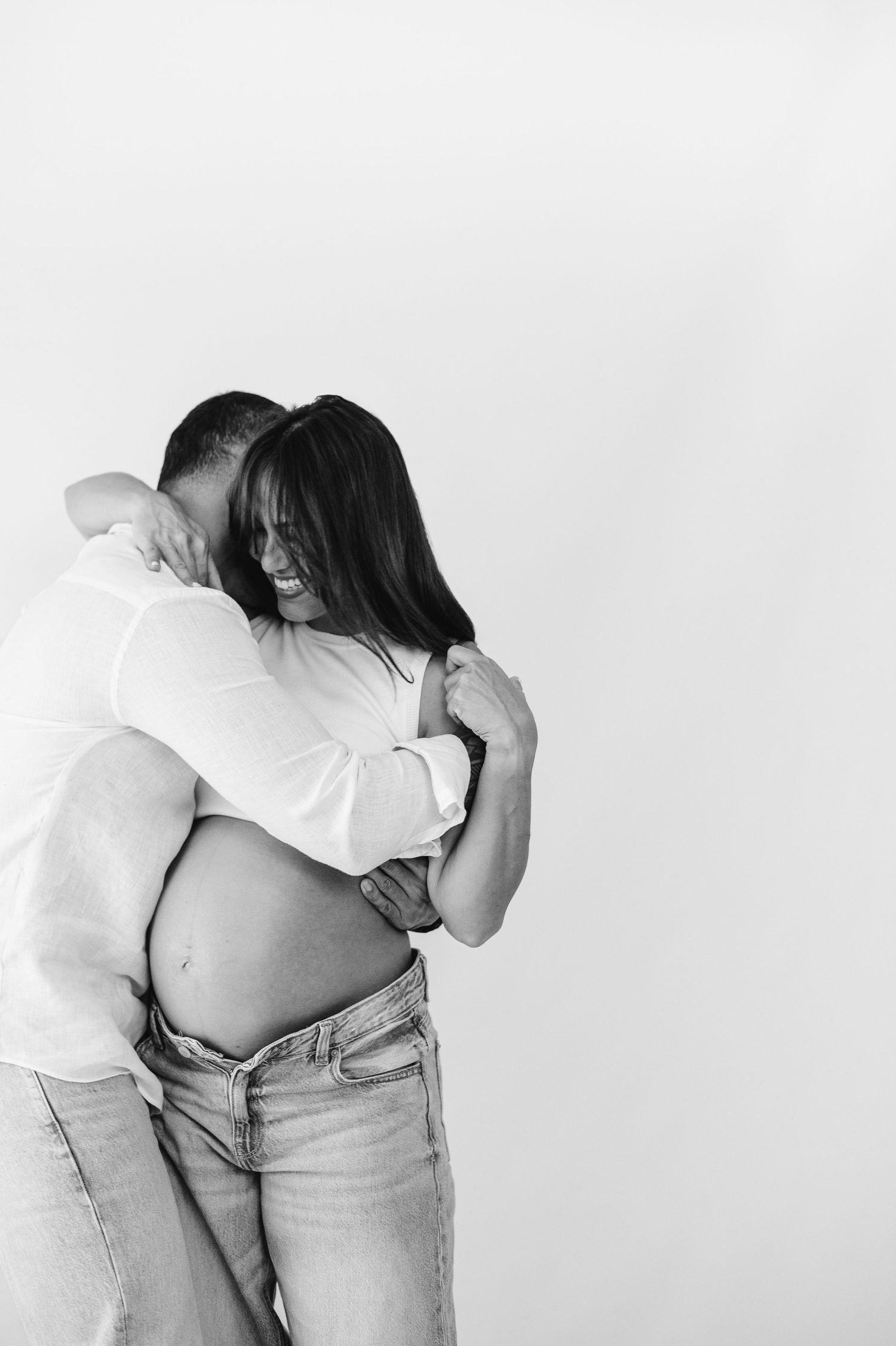 Happy pregnant couple dancing together in white outfits, woman cradling baby bump, man spinning her hand overhead.