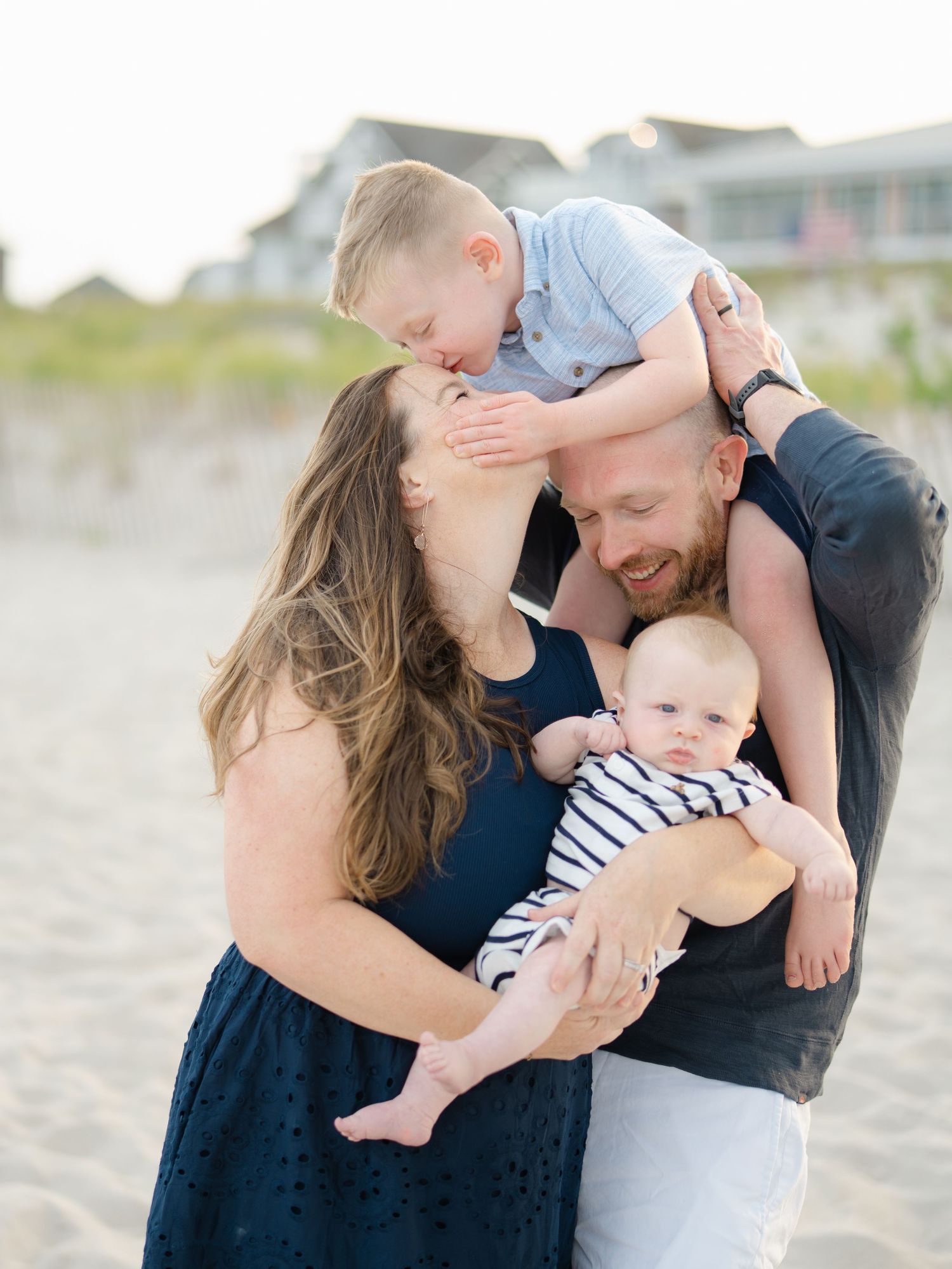 Young family of four snuggle together while son kisses moms head at the Jersey shore beach.