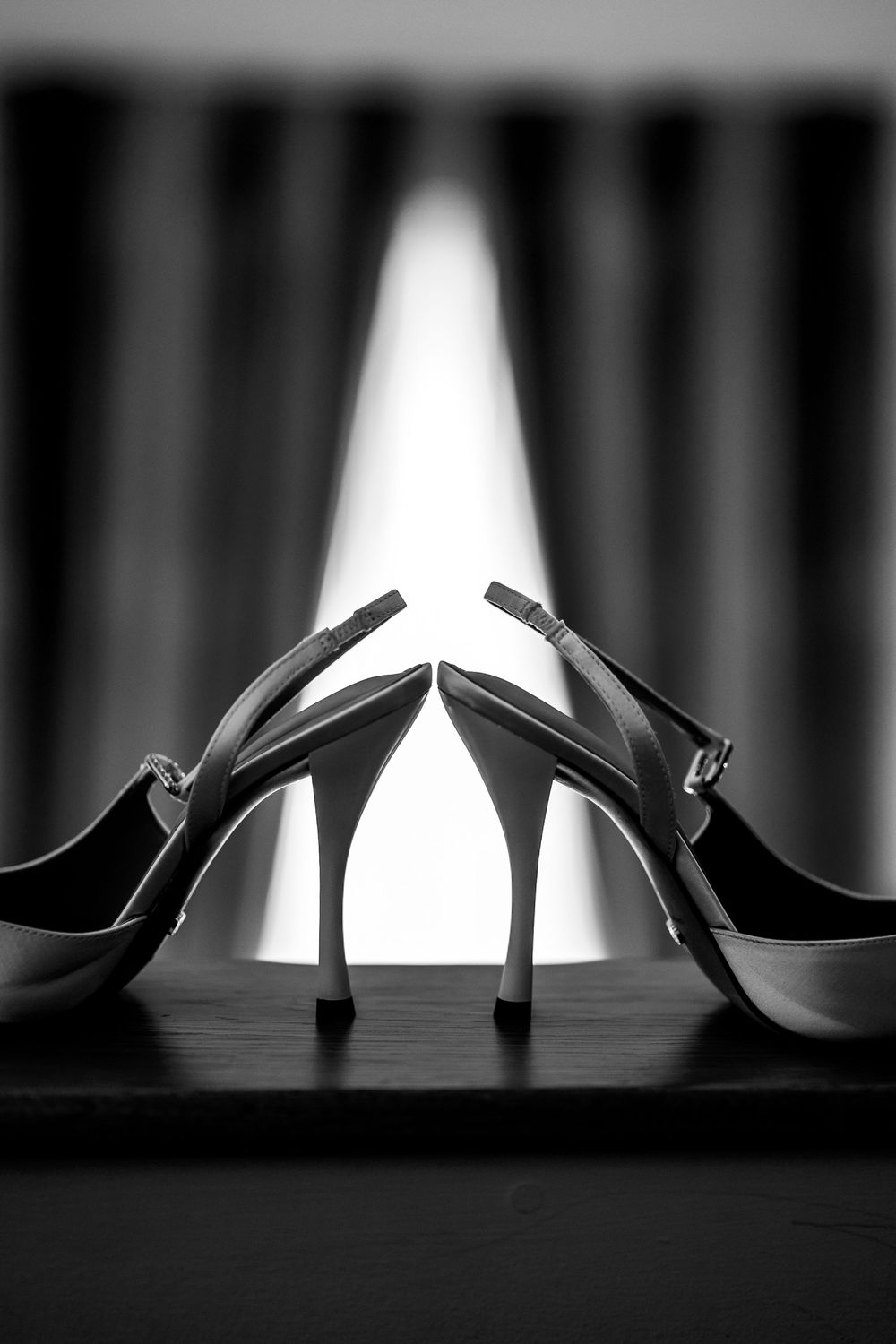 Black and white photo of elegant high heel slingback shoes on a dark surface with dramatic curtain backdrop.