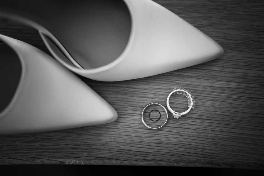 Black and white photo of wedding rings and diamond engagement ring beside elegant white pointed-toe heels on wood surface.