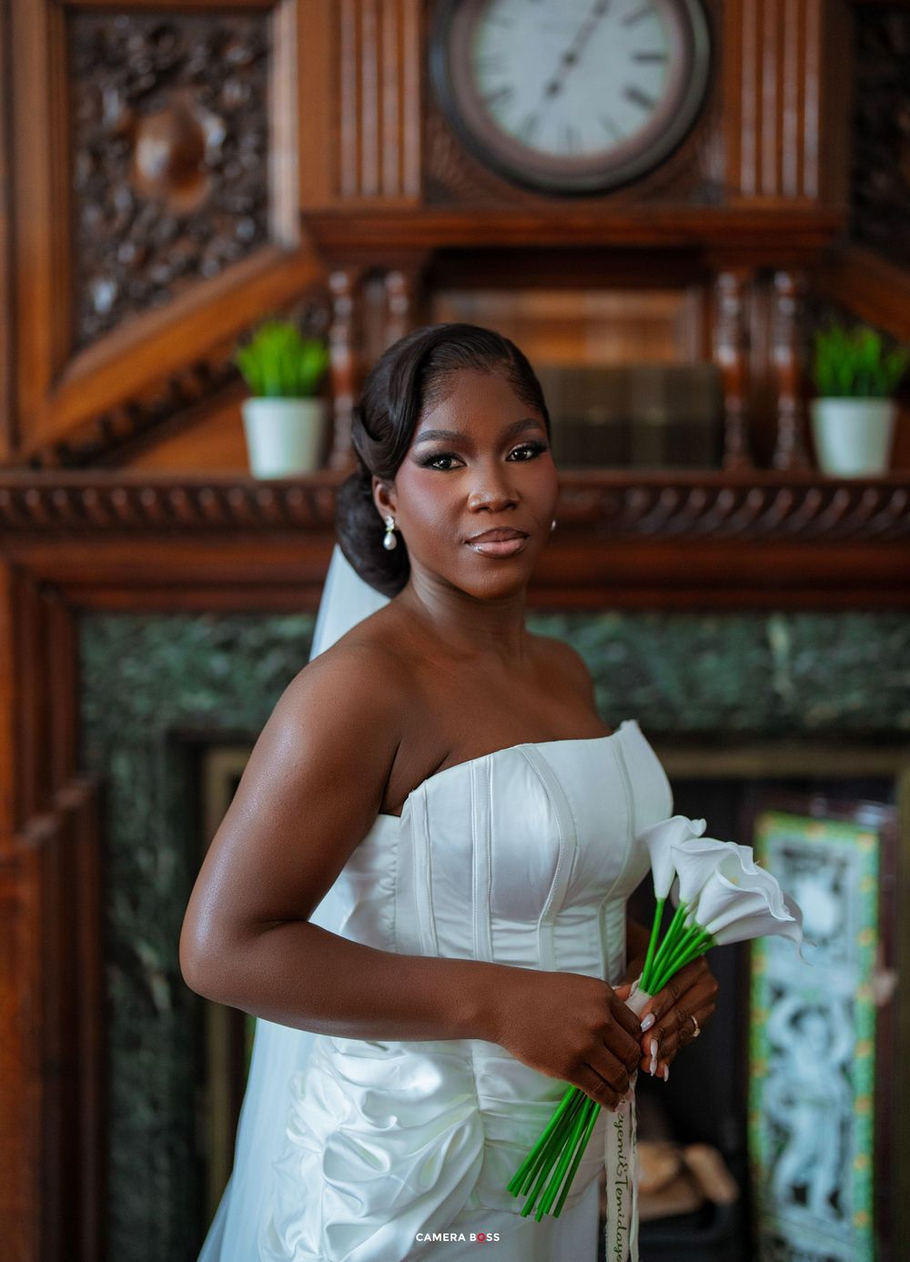 Elegant bride in strapless white gown holding calla lily bouquet, posing before ornate wooden mantel with antique clock.