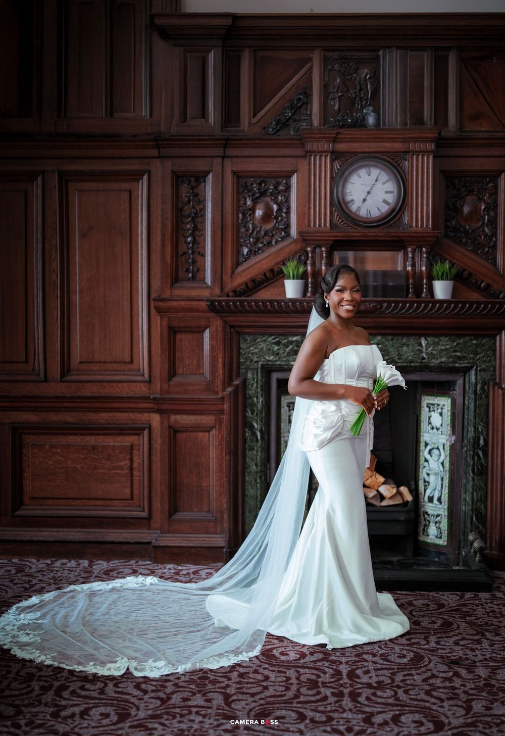 Bride in white strapless gown with long veil poses by ornate fireplace in elegant wood-paneled room.