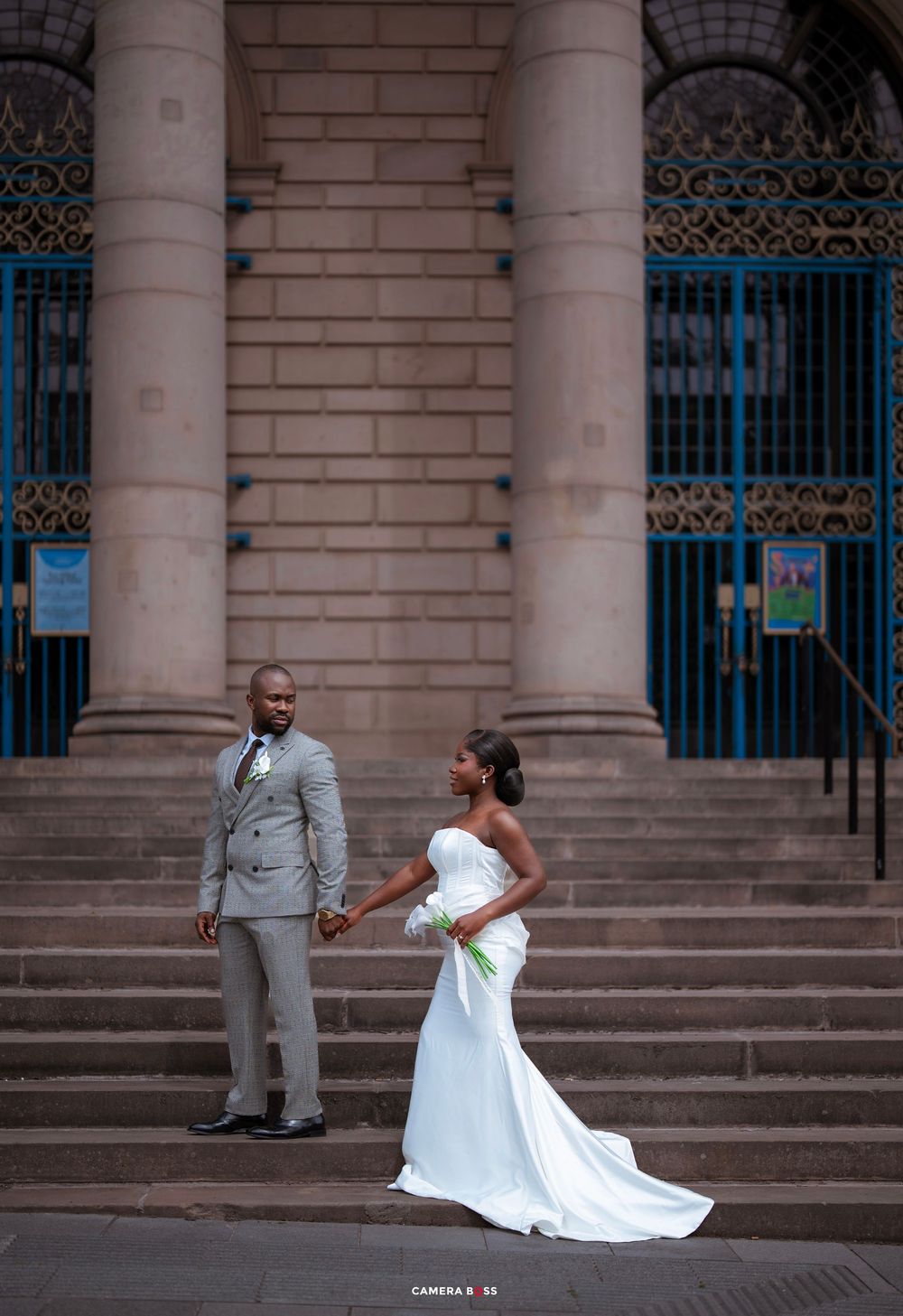 Bride and groom in wedding attire hold hands on steps of grand building with blue gates.