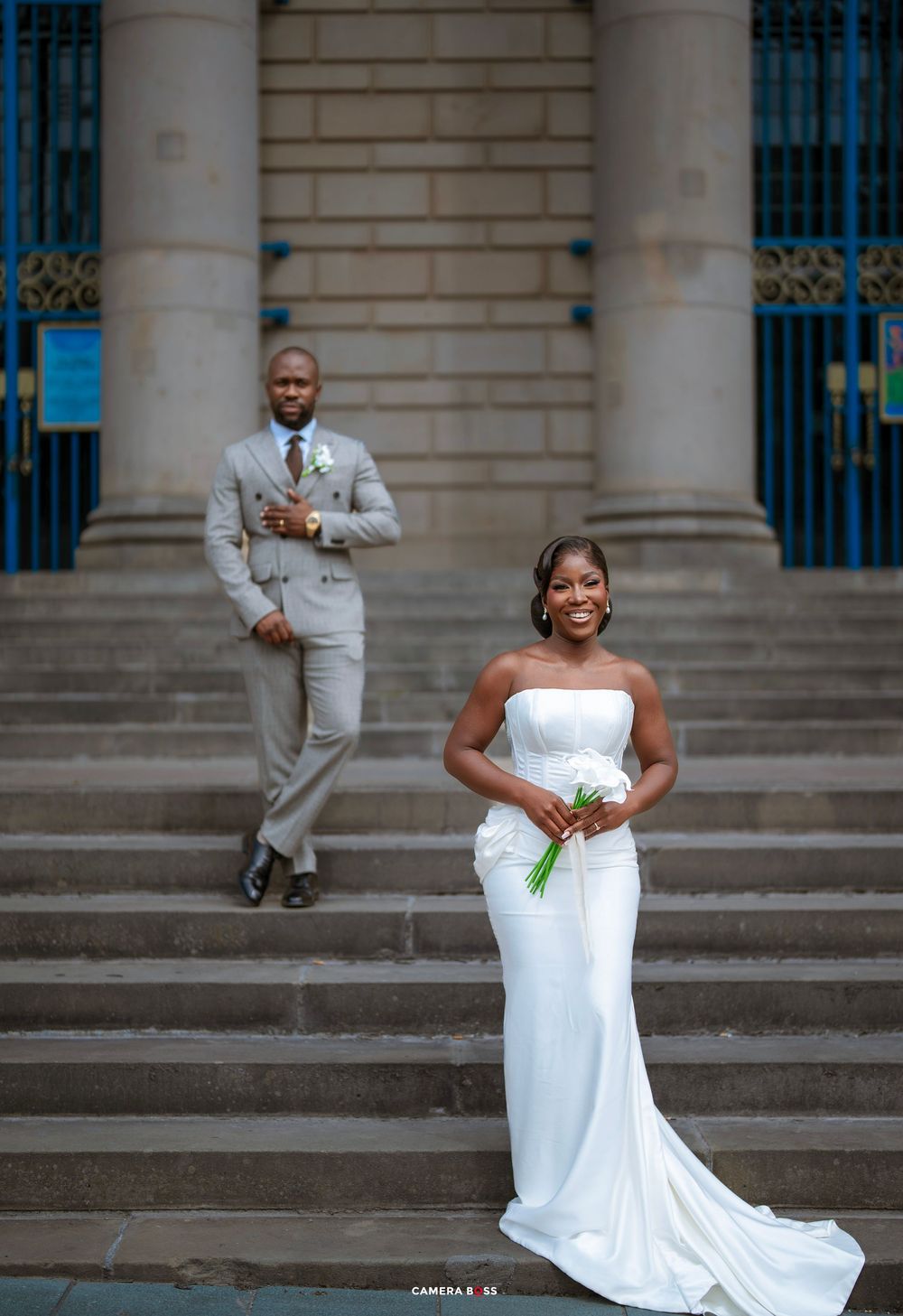 Elegant bride in white strapless gown holds bouquet on steps as groom in gray suit stands behind at wedding venue.