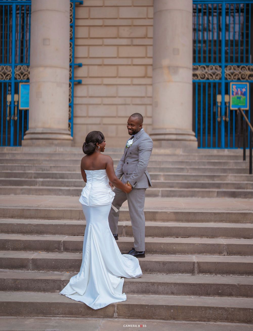 A bride in a white mermaid gown and groom in gray suit hold hands on stone steps outside a grand building.