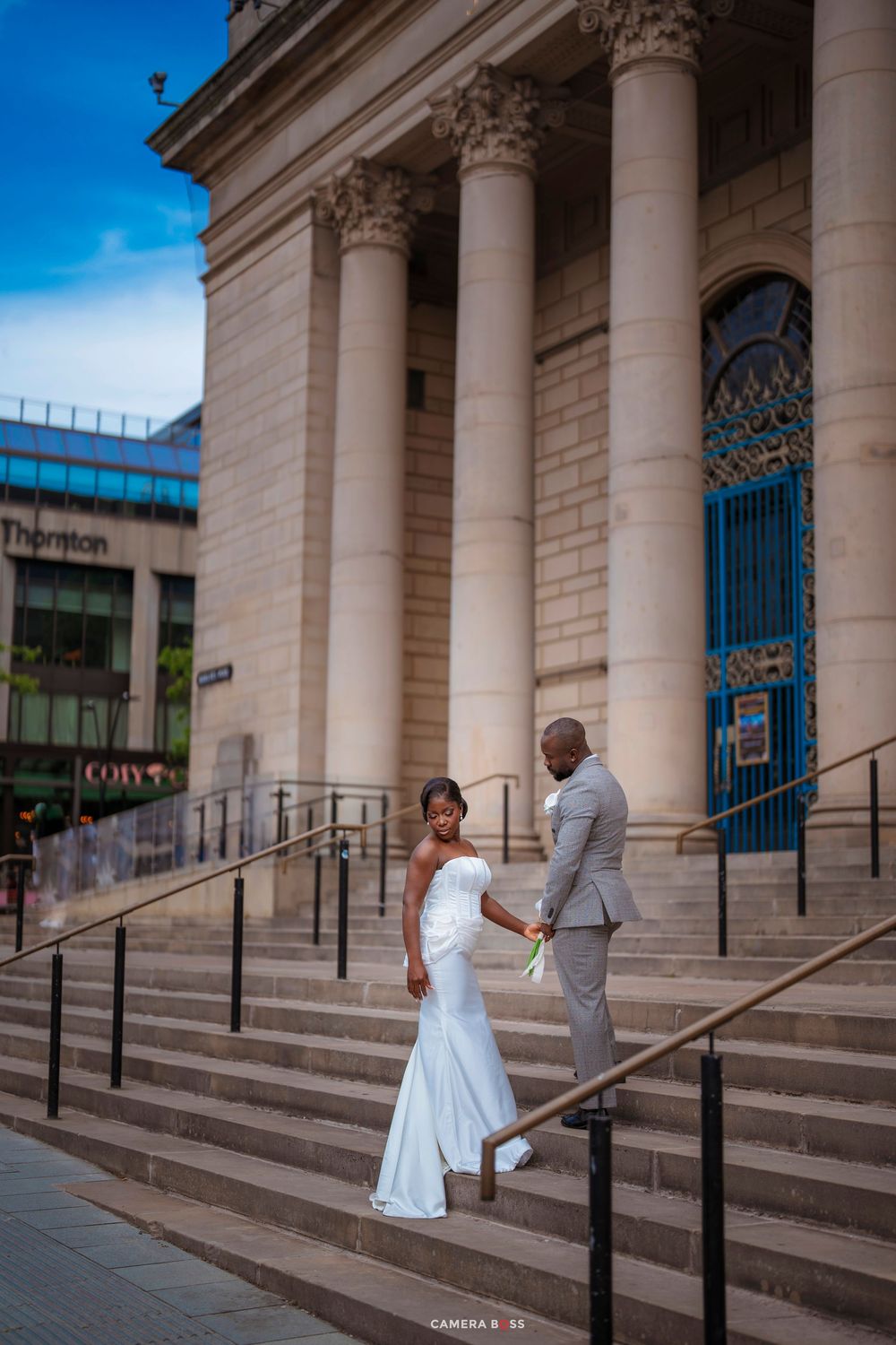 Bride in white gown and groom in gray suit pose on steps of grand neoclassical building on their wedding day.
