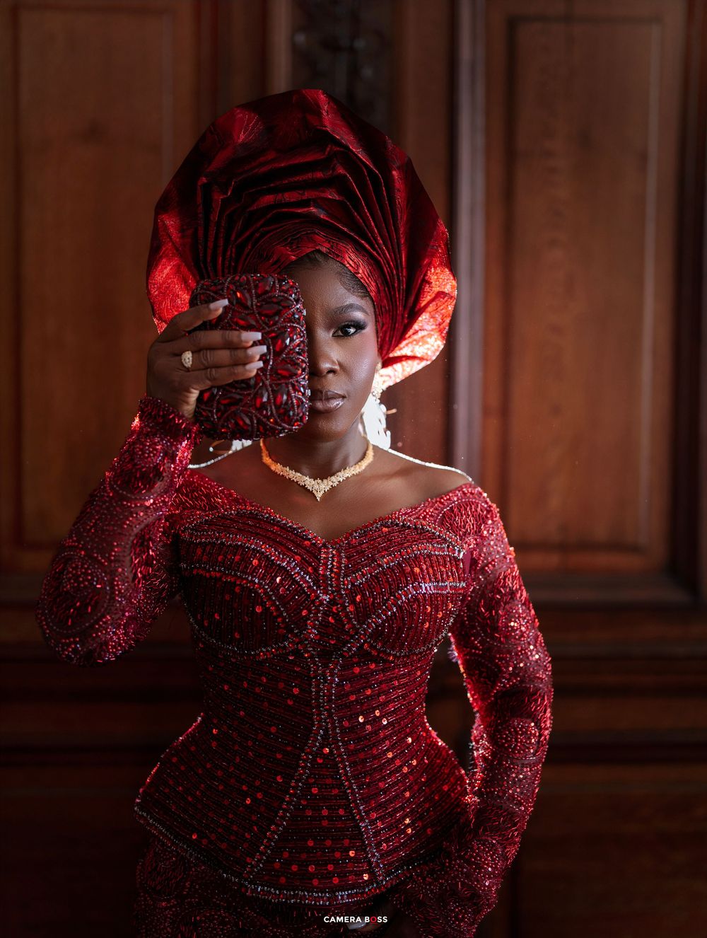 Woman in red beaded Nigerian traditional attire with gele headwrap holds clutch purse elegantly.