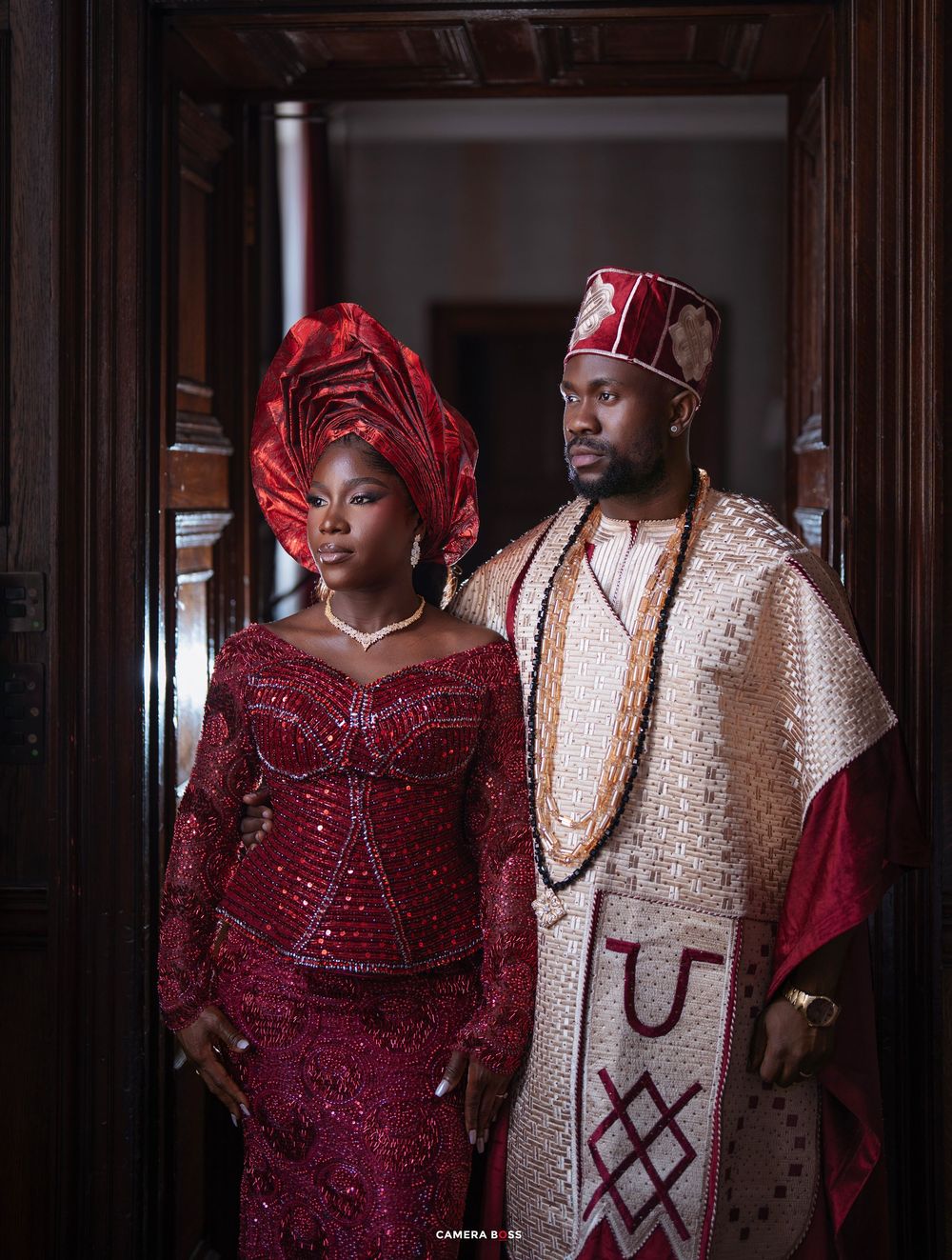 Couple in elegant Nigerian traditional attire, woman in red beaded gele and gown, man in embroidered agbada with cap.