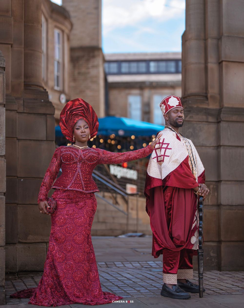 Couple in traditional Nigerian attire, woman in red lace gown and gele, man in red and white agbada, posing outdoors.