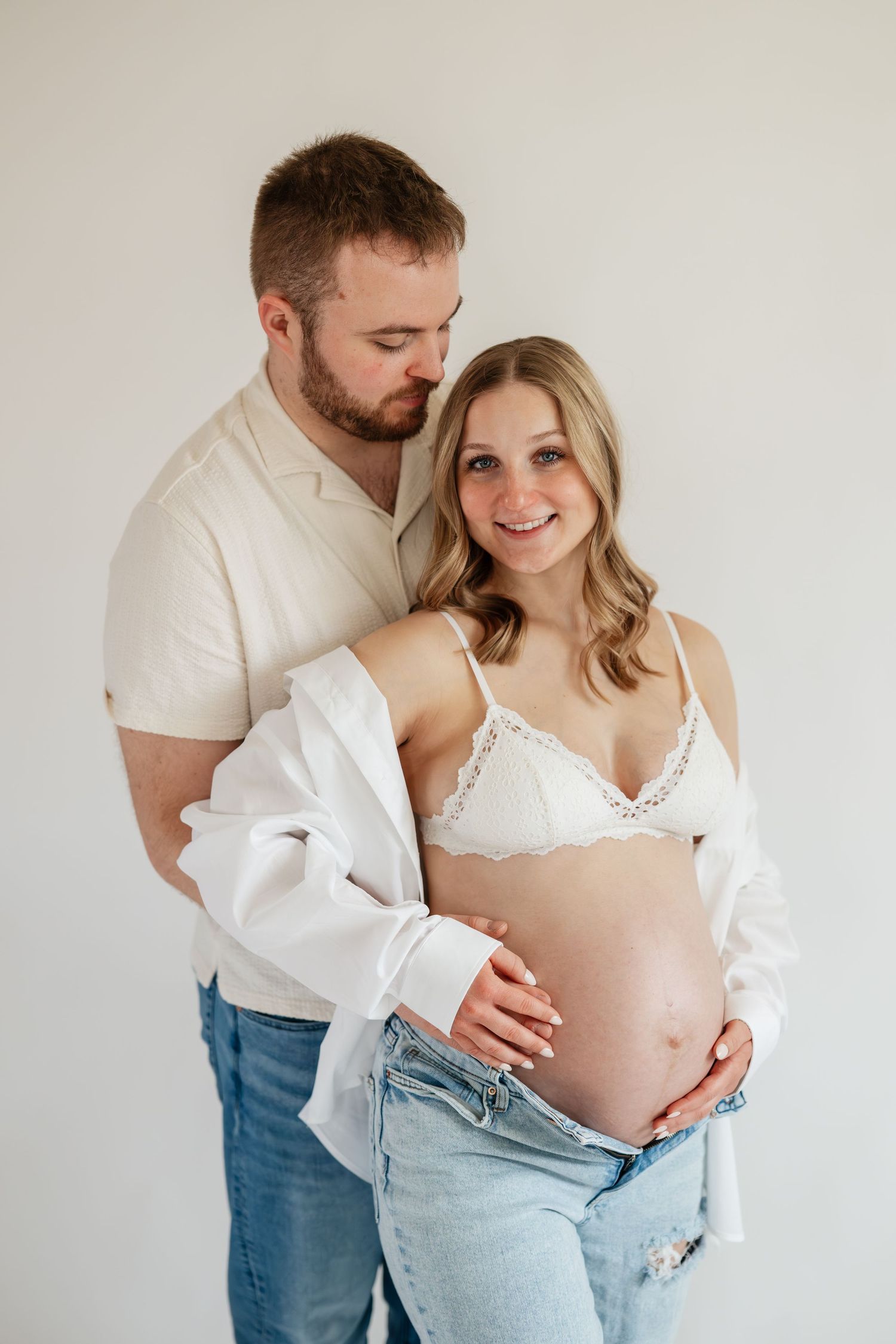 Pregnant woman in ruffled off-shoulder dress cradling baby bump, lying on soft bedding.