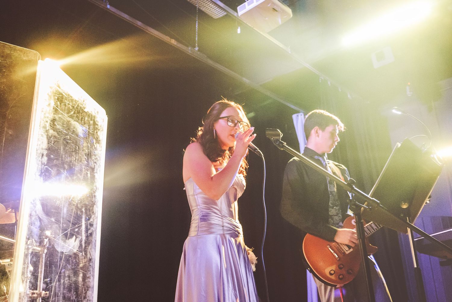 A woman in a silver gown sings into a microphone on a lit stage while a guitarist performs behind her.