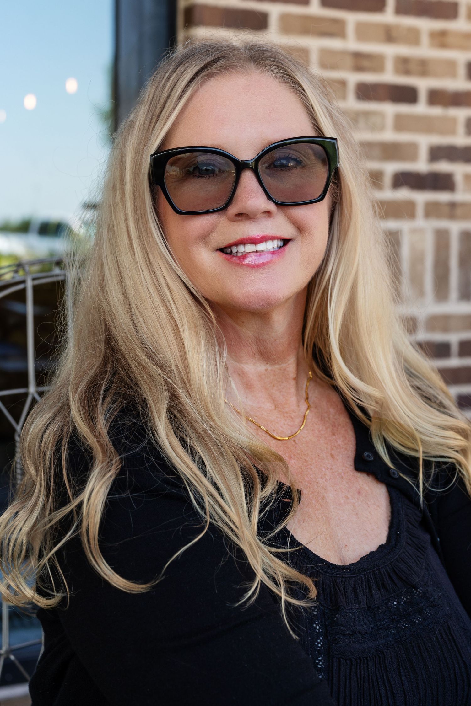Blonde woman wearing black sunglasses and black top smiling outdoors near brick wall.