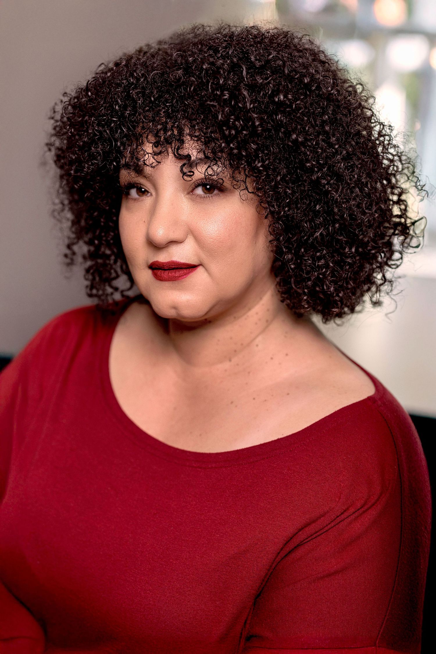 Woman with curly dark hair wearing a red top and red lipstick poses for a portrait photo.