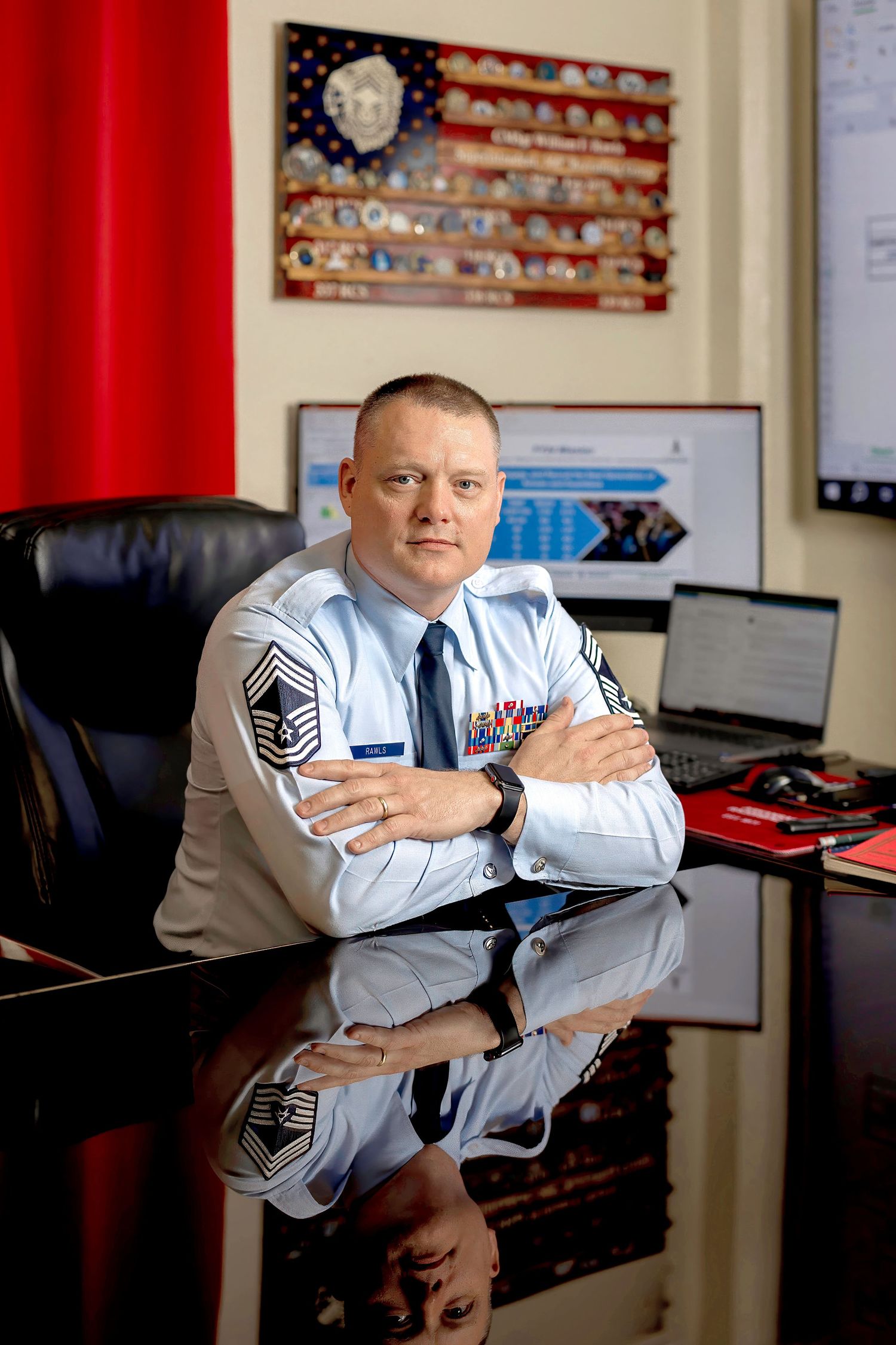 Senior military officer in dress uniform sits at office desk with arms crossed, US flag display and computer visible behind him.