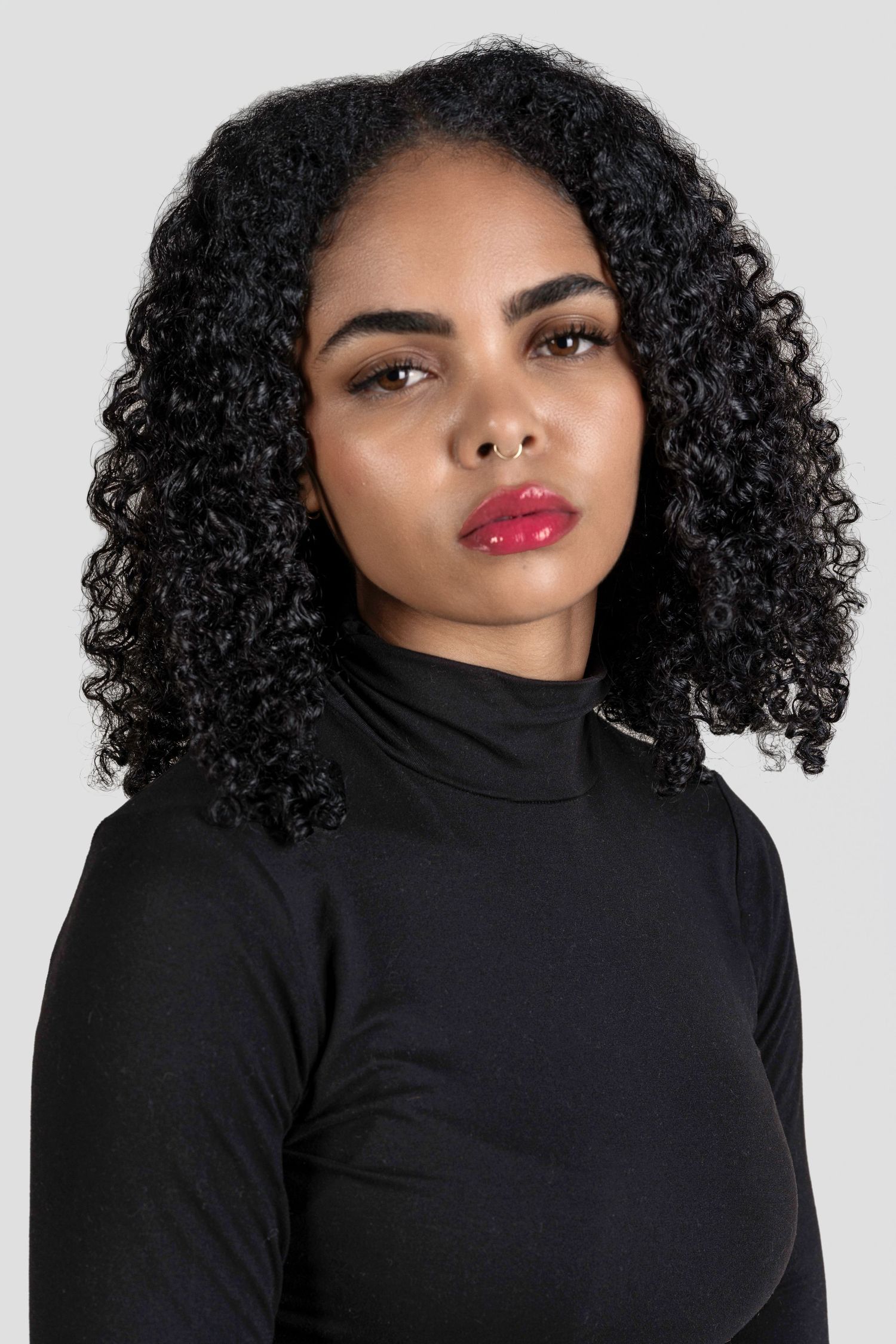 Young Black woman with curly hair wearing a black turtleneck, red lipstick, posing against a gray background.