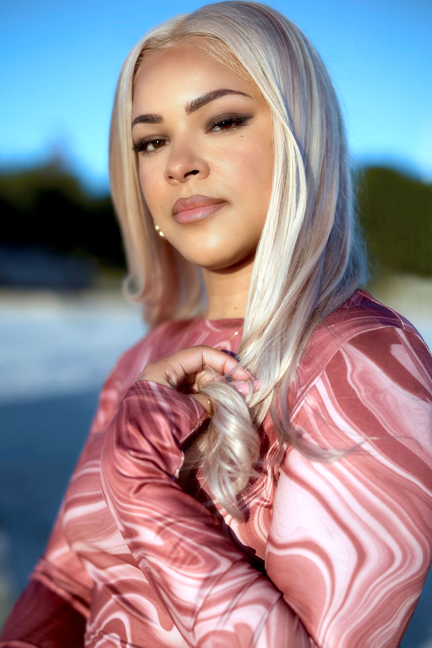 Woman with platinum blonde hair wearing a pink marble-print top poses outdoors against a blue sky backdrop.