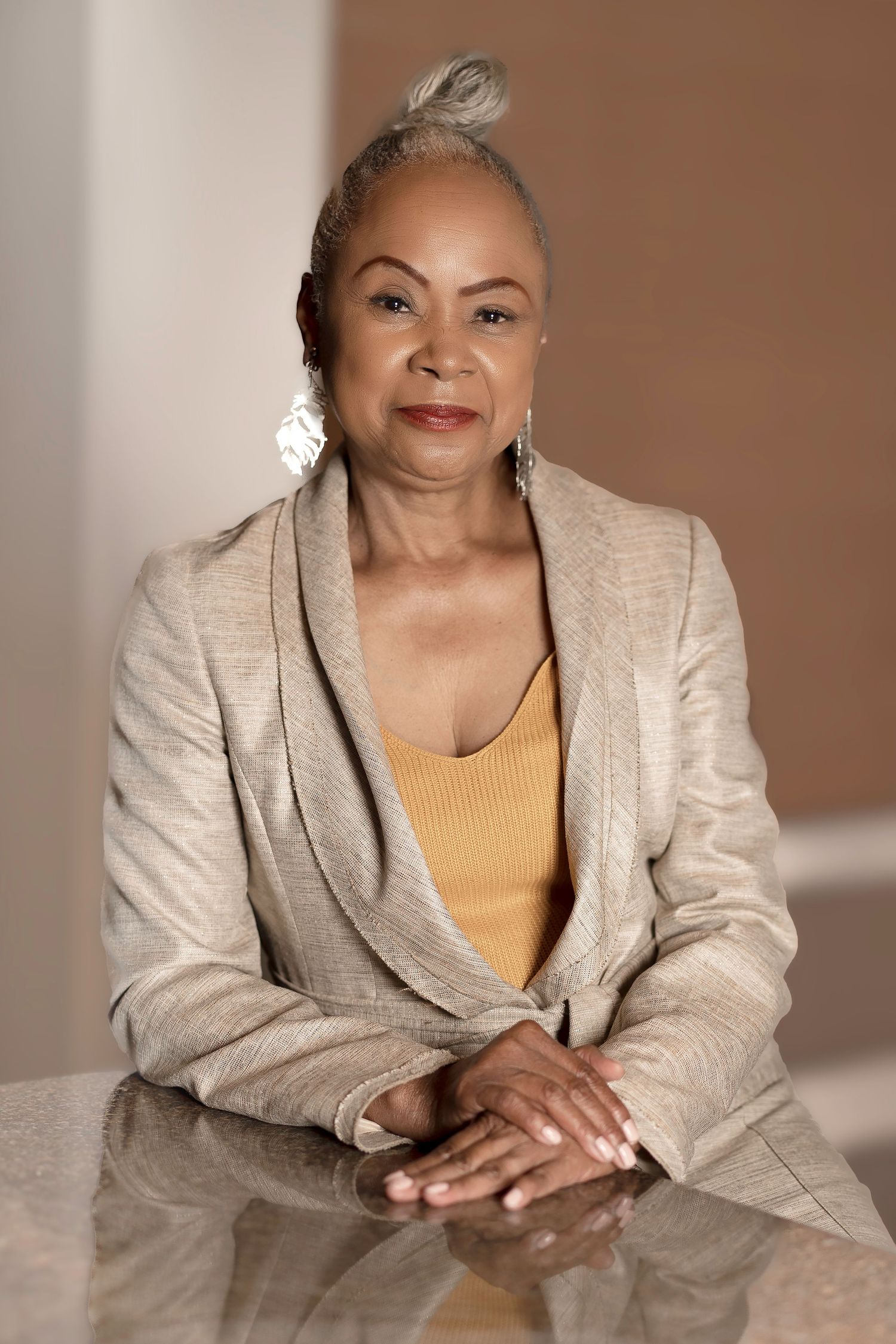 Elegant older Black woman in beige blazer and gold top, sitting confidently at a table with hands folded, smiling.