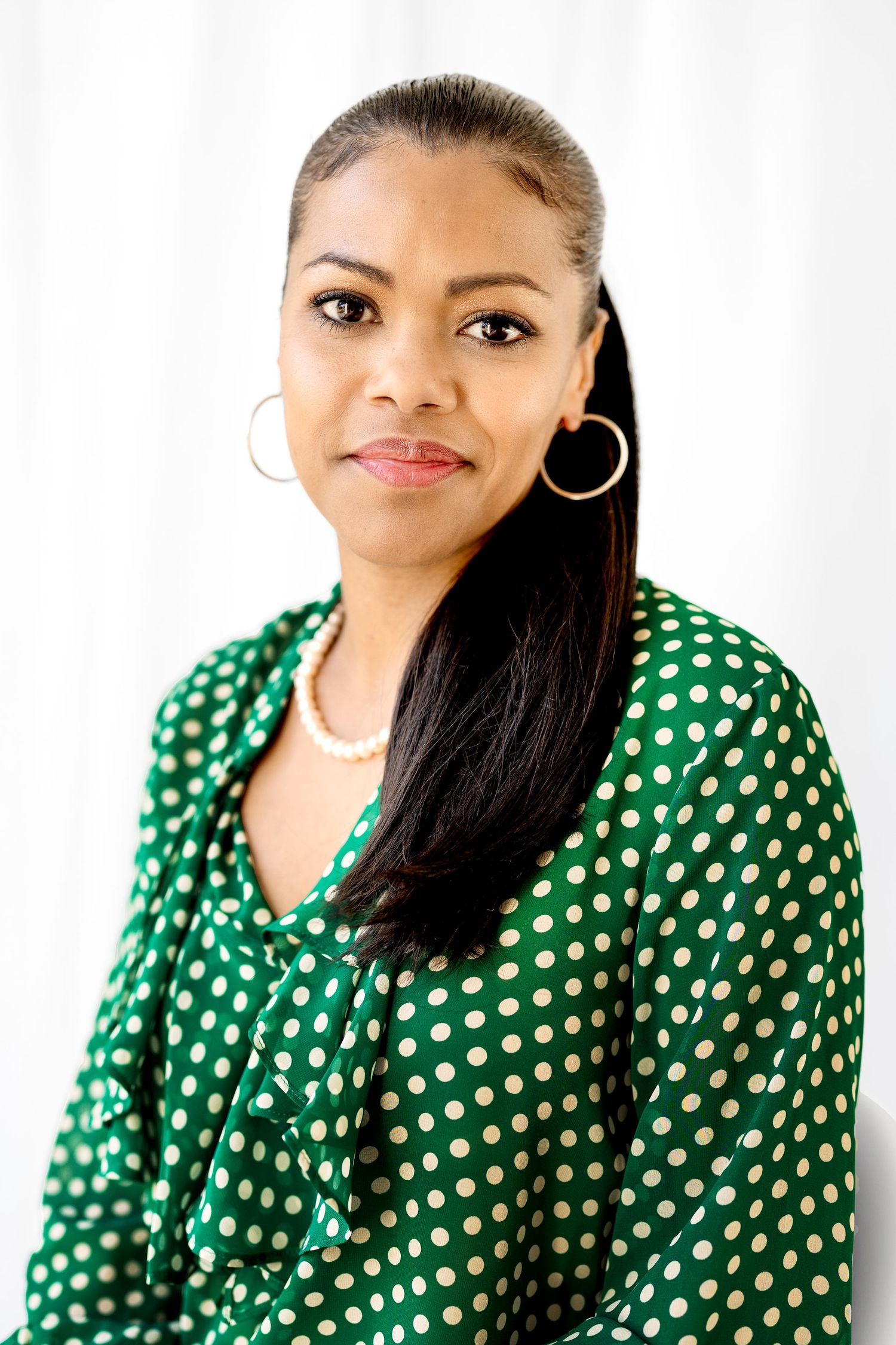 Woman in green polka dot blouse with hoop earrings and pearl necklace smiling at camera.