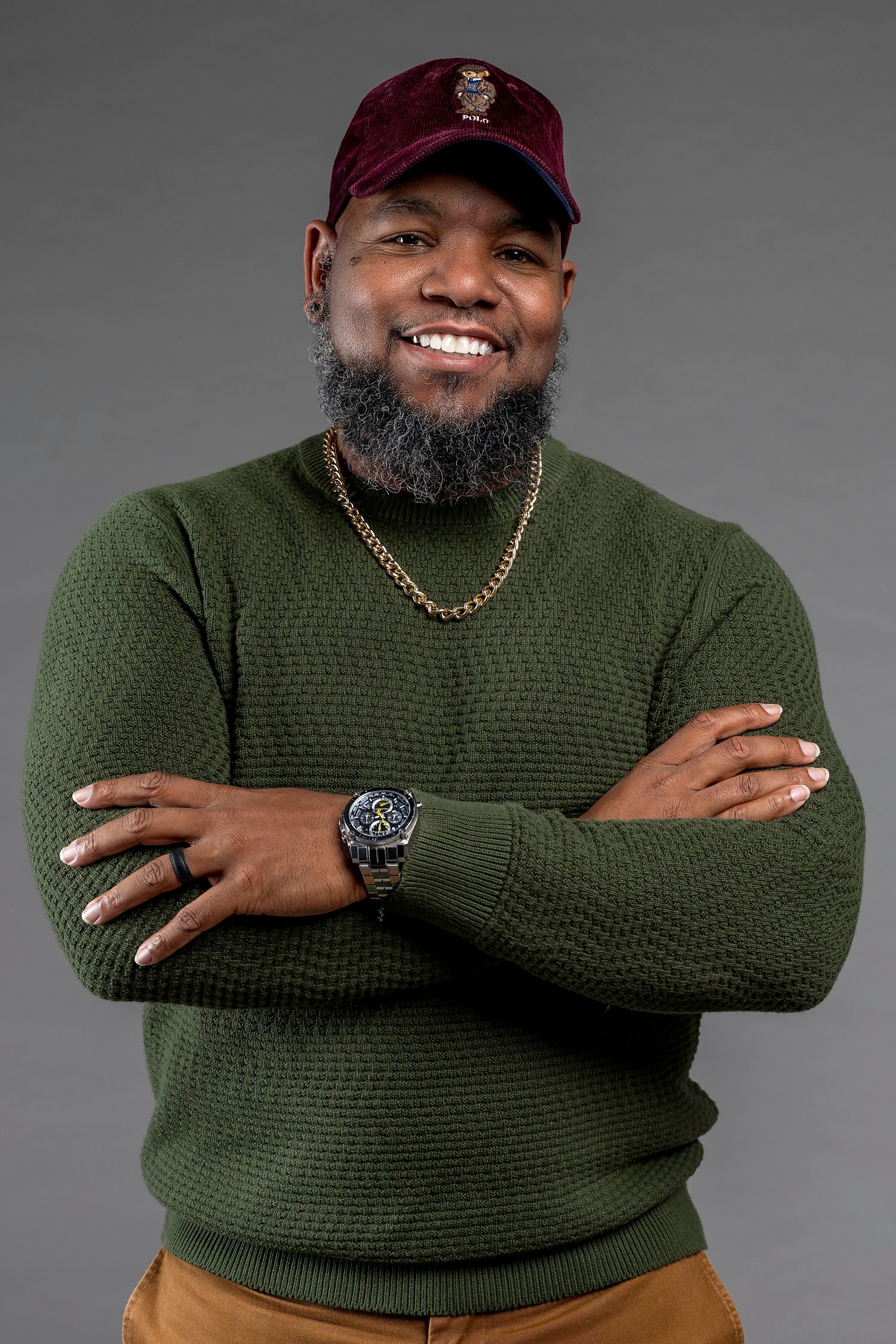 Smiling bearded Black man in green sweater, burgundy cap, gold chain, and watch, posing with arms crossed.