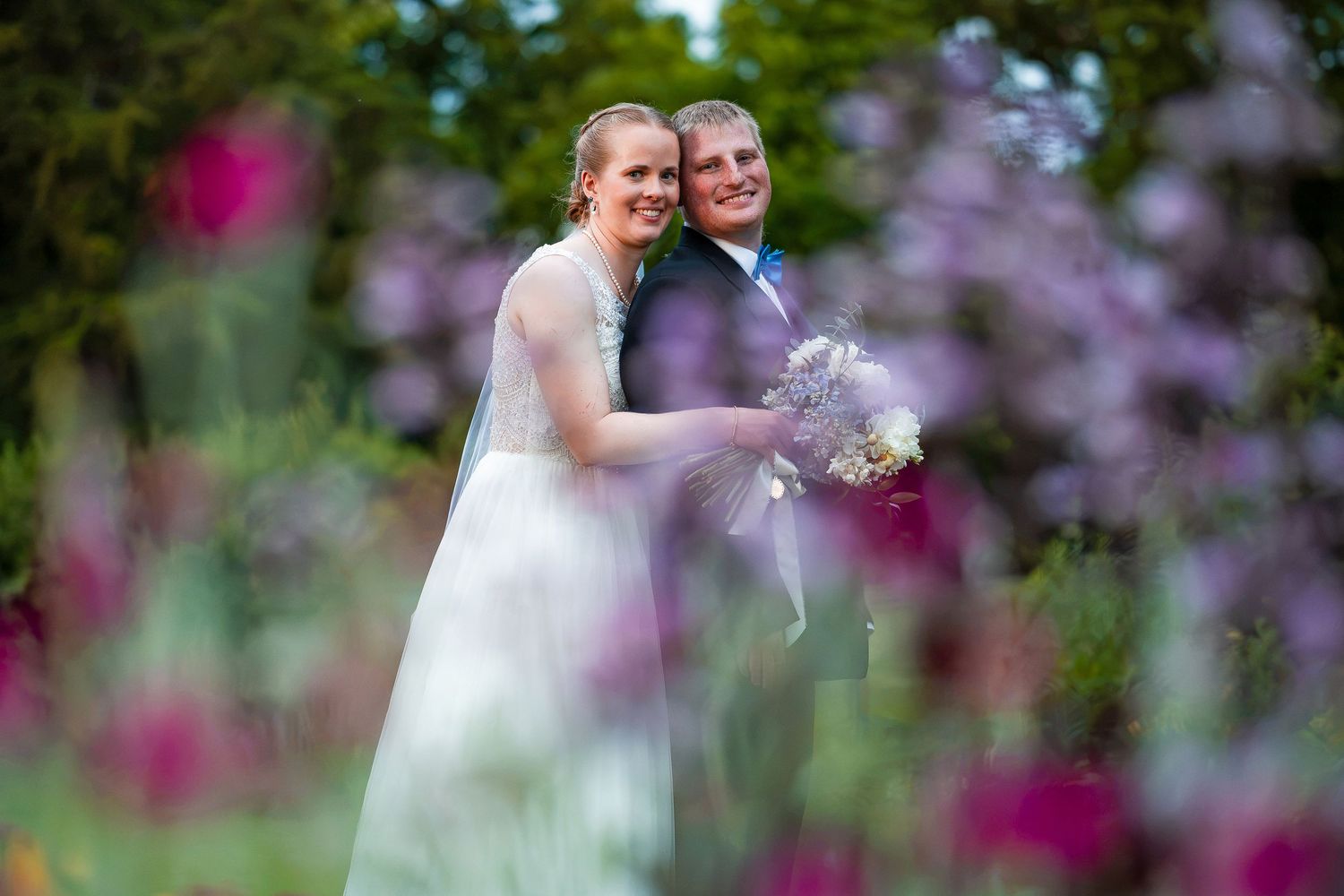 Bride and groom smiling in a lush garden surrounded by vibrant pink and purple flowers on their wedding day.