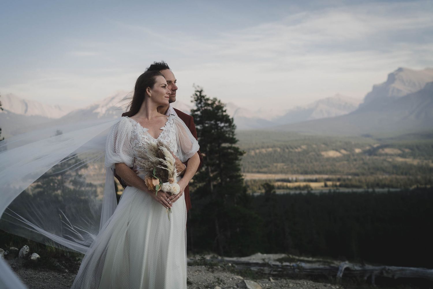 Bride in white gown with flowing veil holds bouquet on mountain overlook with scenic Rocky Mountain backdrop.