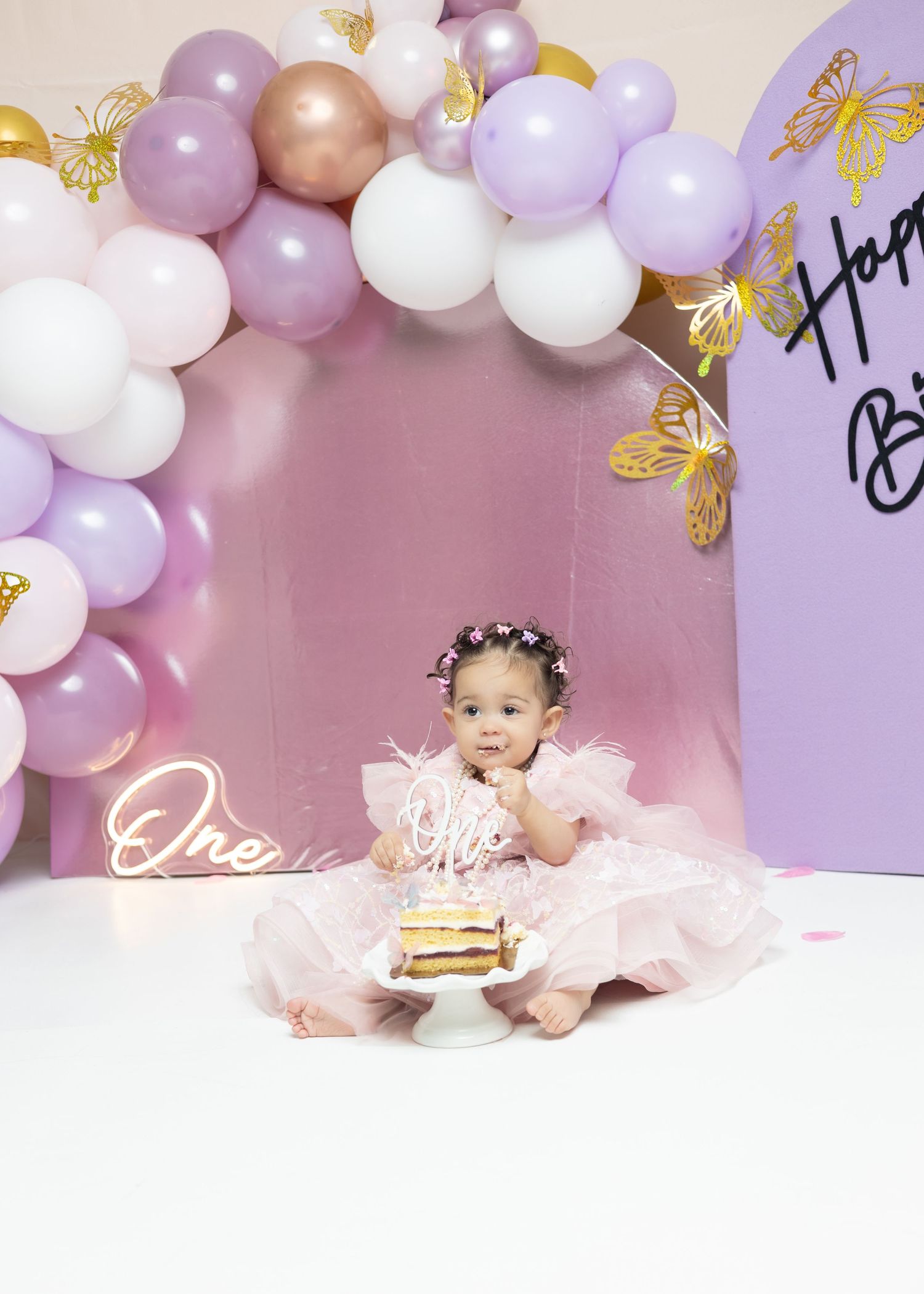 Baby girl in pink tutu sits before first birthday cake surrounded by purple and white balloon arch with gold butterflies.