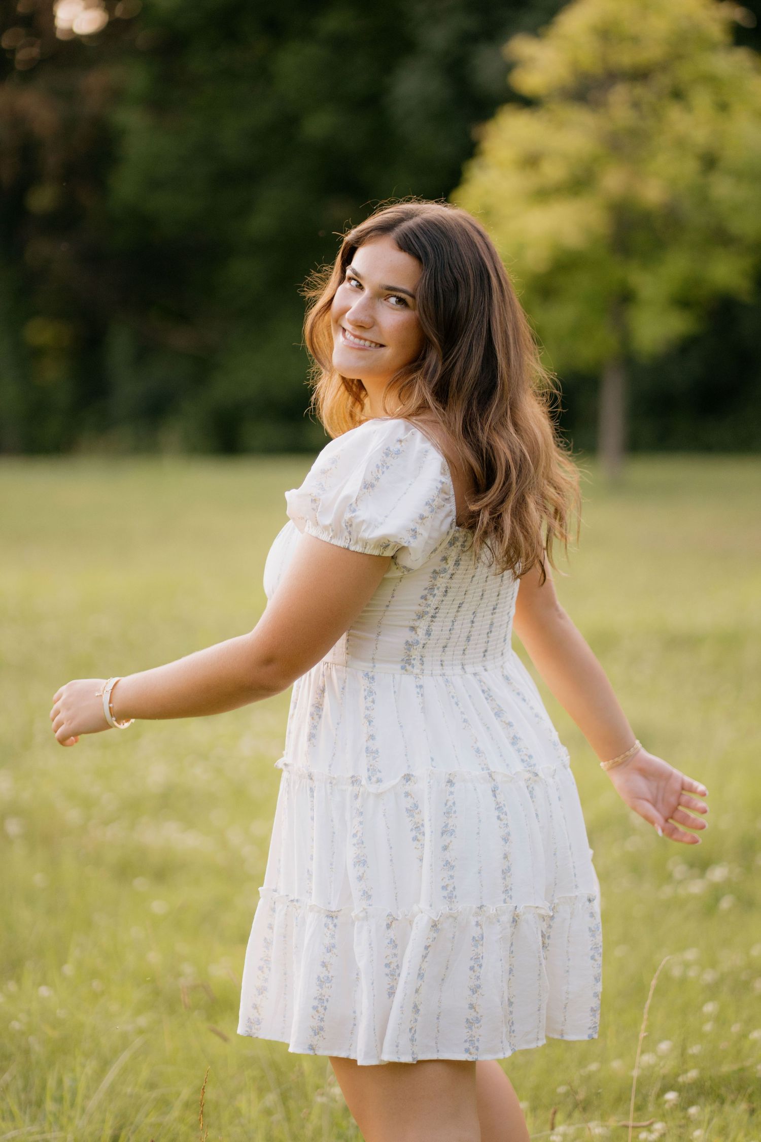 Woman in white polka dot sundress standing in green field with trees in background on sunny day.