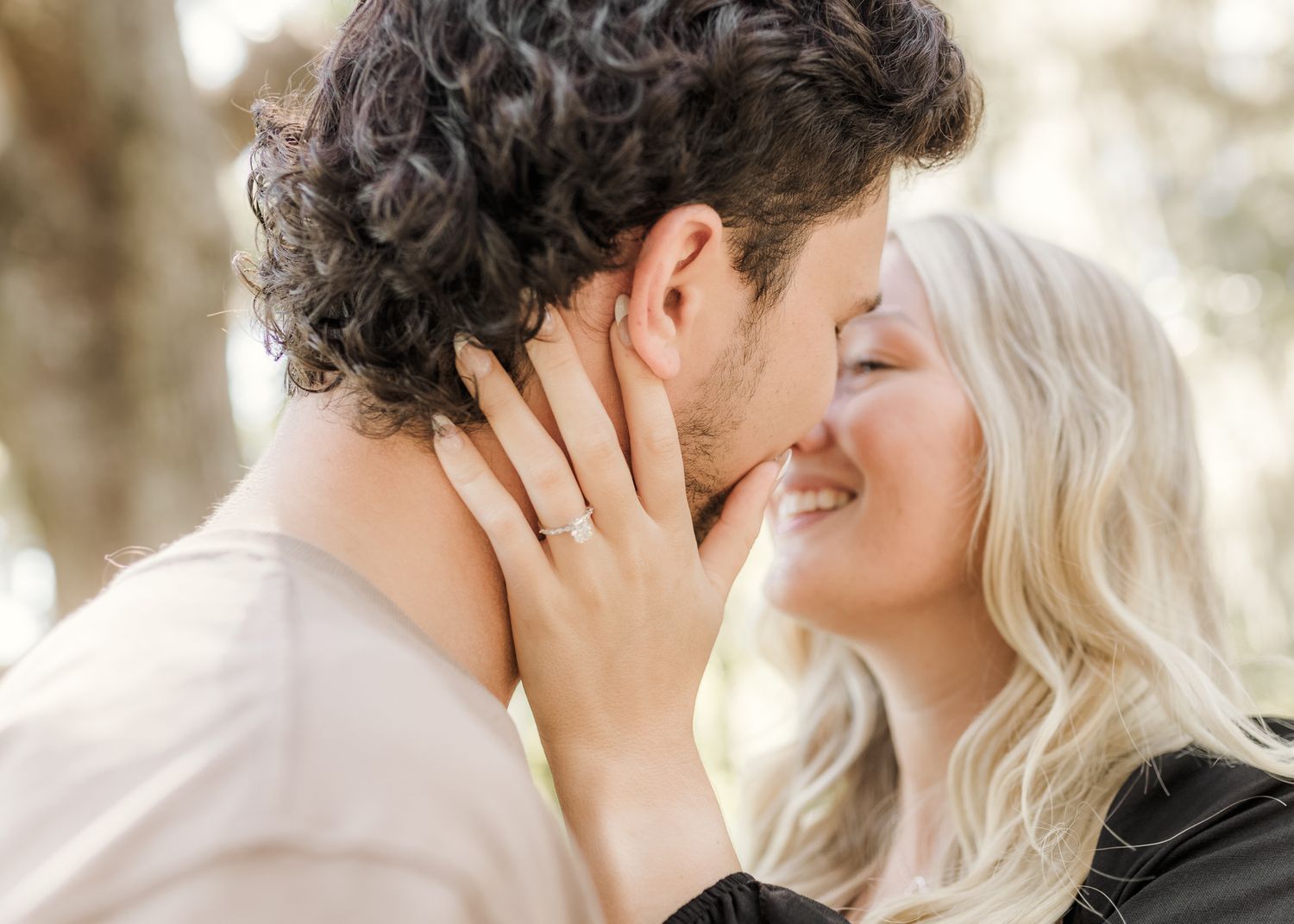 A couple shares a romantic kiss outdoors, the woman smiling and showing off her engagement ring.