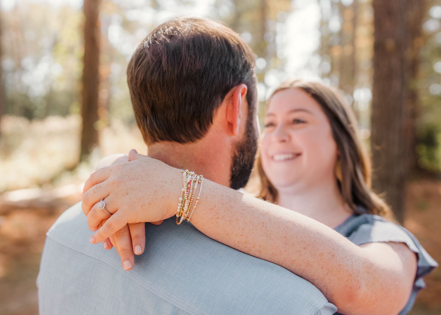 A smiling woman embraces a bearded man outdoors, wearing gold stacked bracelets and a ring, with sunlit trees in the background.