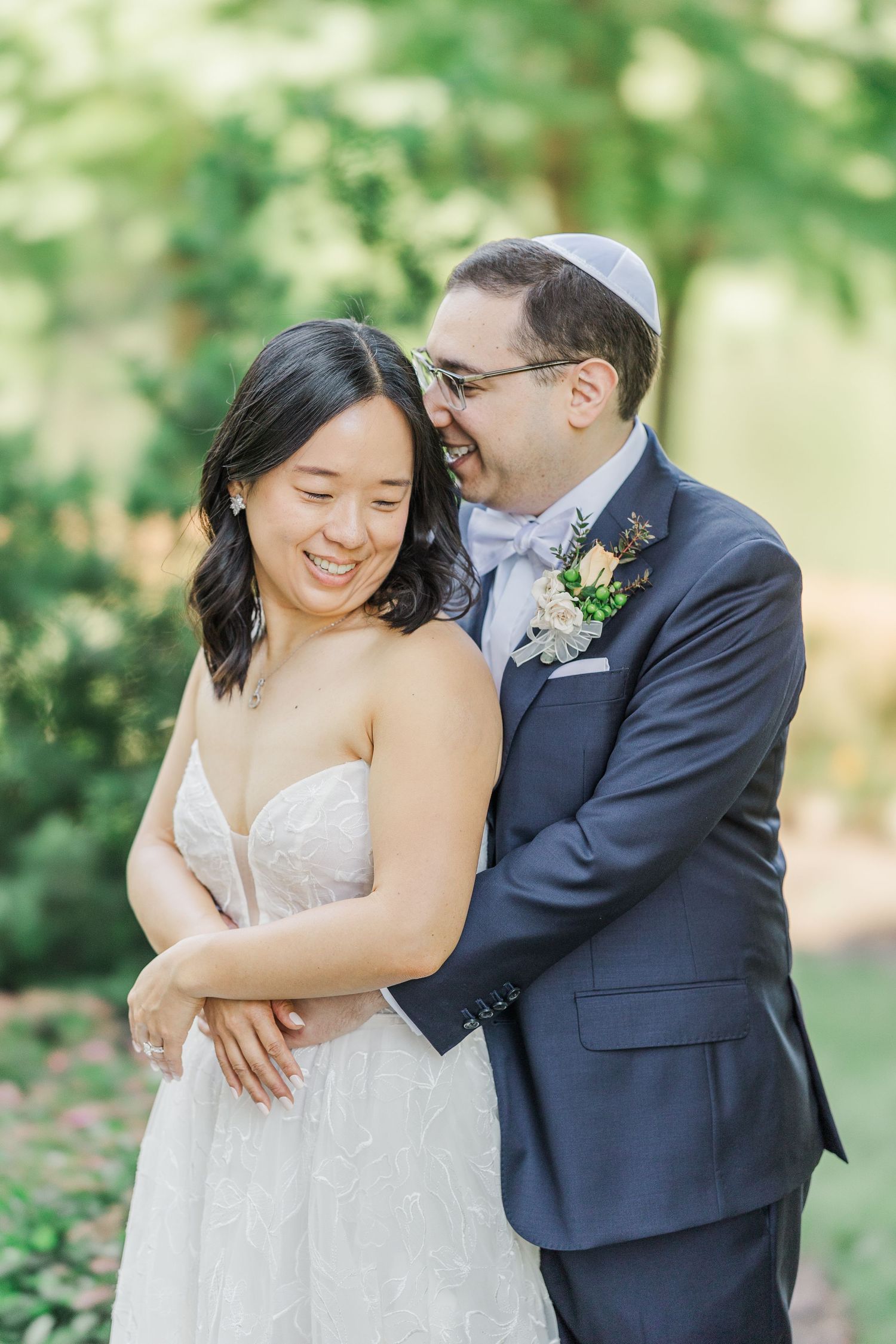 Joyful bride and groom embracing outdoors, she in lace gown, he in navy suit with kippah and floral boutonniere.