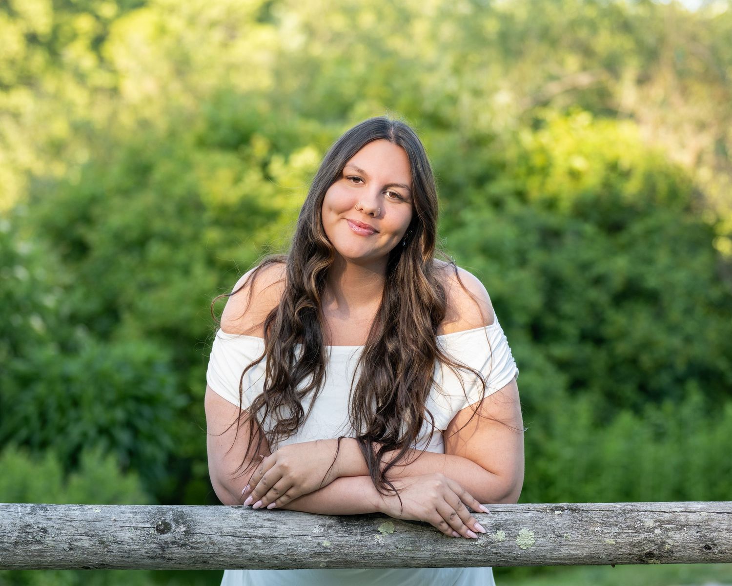 Young woman with long brown hair smiles leaning on a wooden fence, surrounded by lush green trees outdoors.