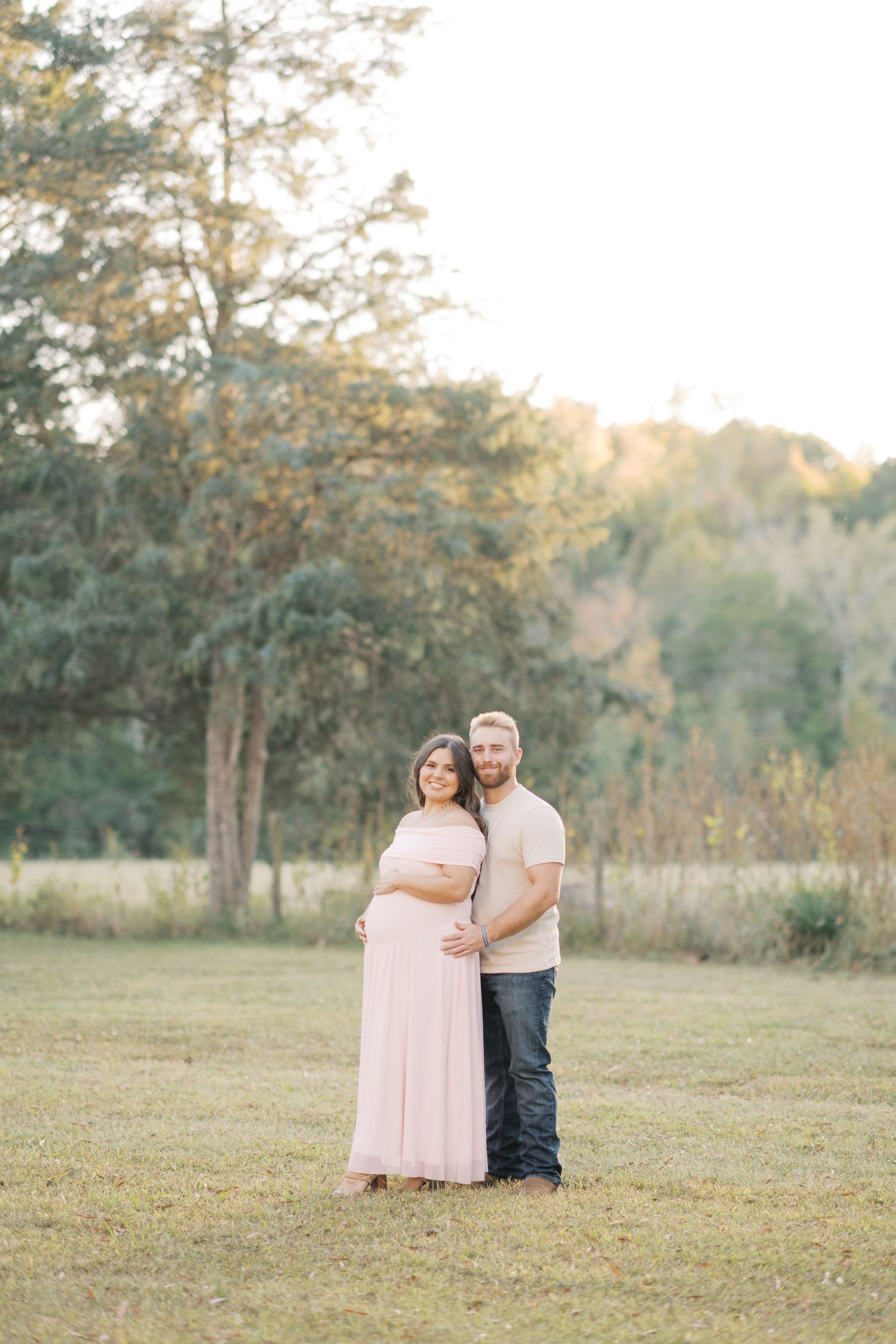 Pregnant couple posing outdoors in golden hour light, woman in pink maternity gown, man in beige shirt and jeans.