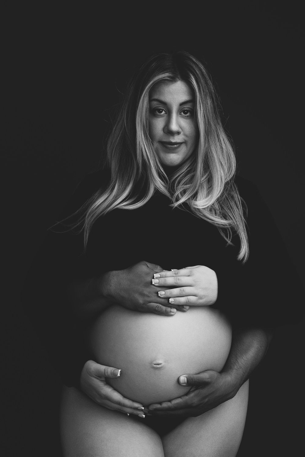 Black and white maternity photo of a blonde woman cradling her pregnant belly against a dark background.