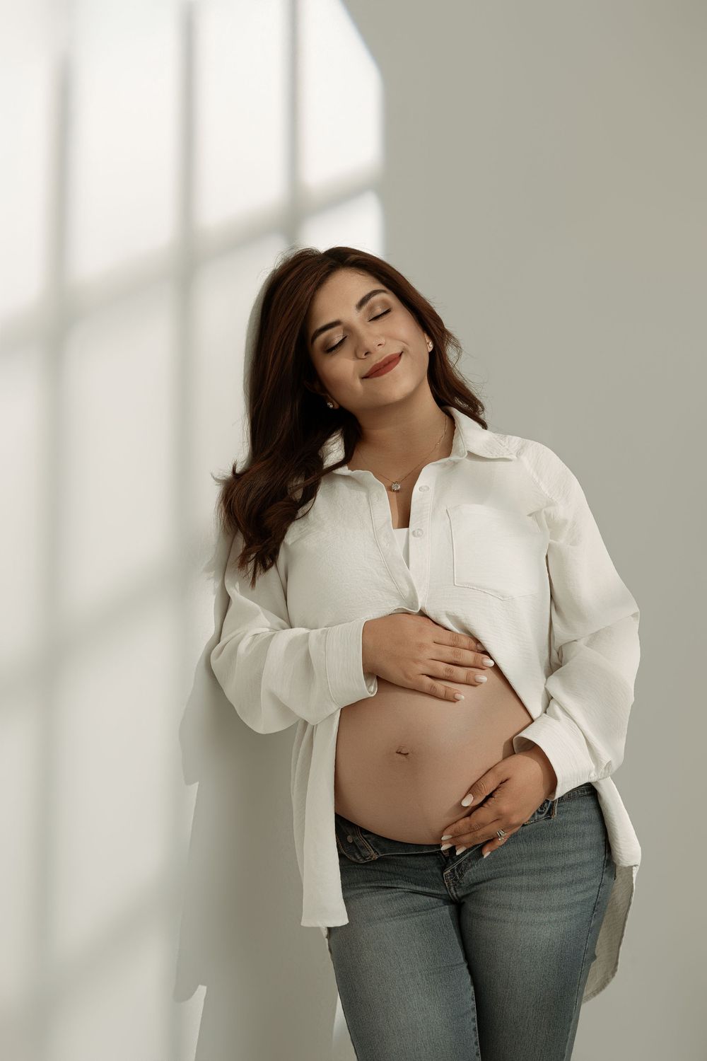 Pregnant woman in white shirt and jeans cradling her baby bump, eyes closed, smiling serenely against a white wall.