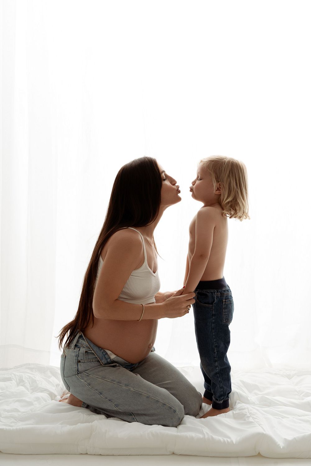 Pregnant mother kneeling on bed sharing a kiss with her young toddler child in a bright white room.