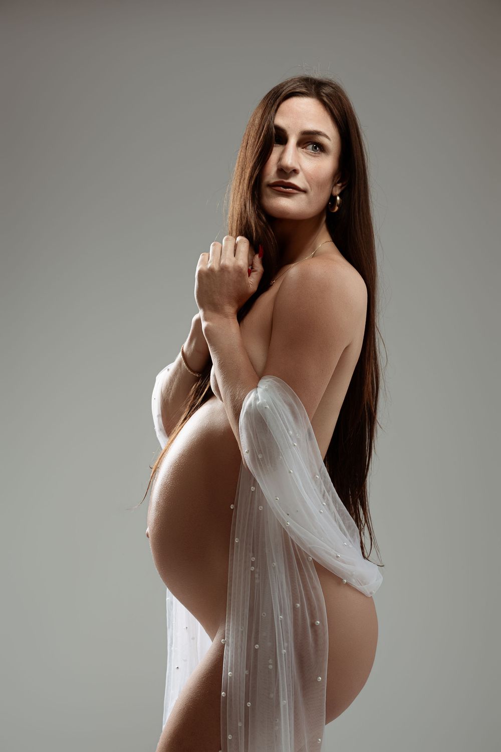 Pregnant woman draped in sheer white pearl-adorned fabric posing gracefully against a neutral gray studio background.