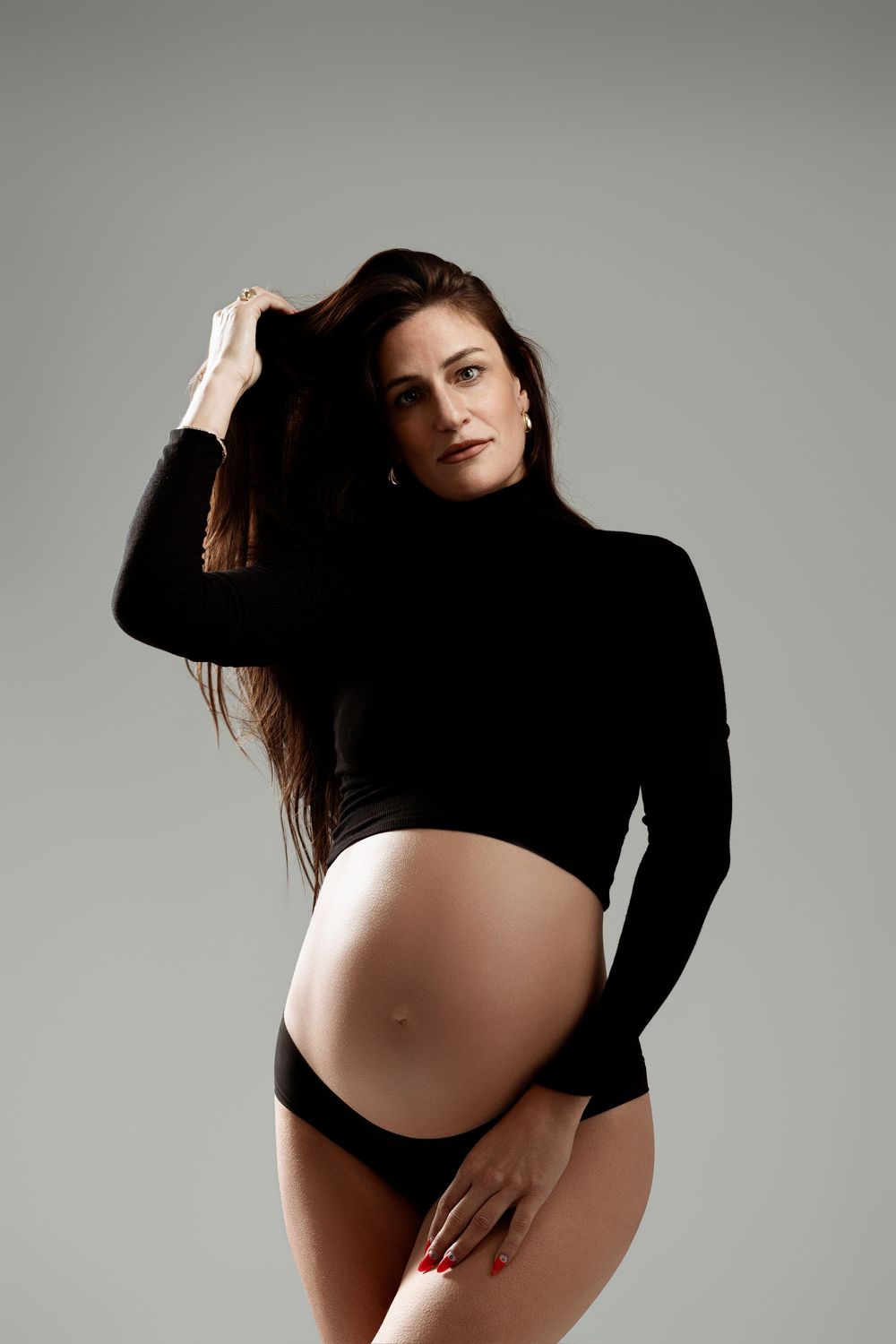 Pregnant woman in black turtleneck crop top and underwear posing gracefully in a studio setting.