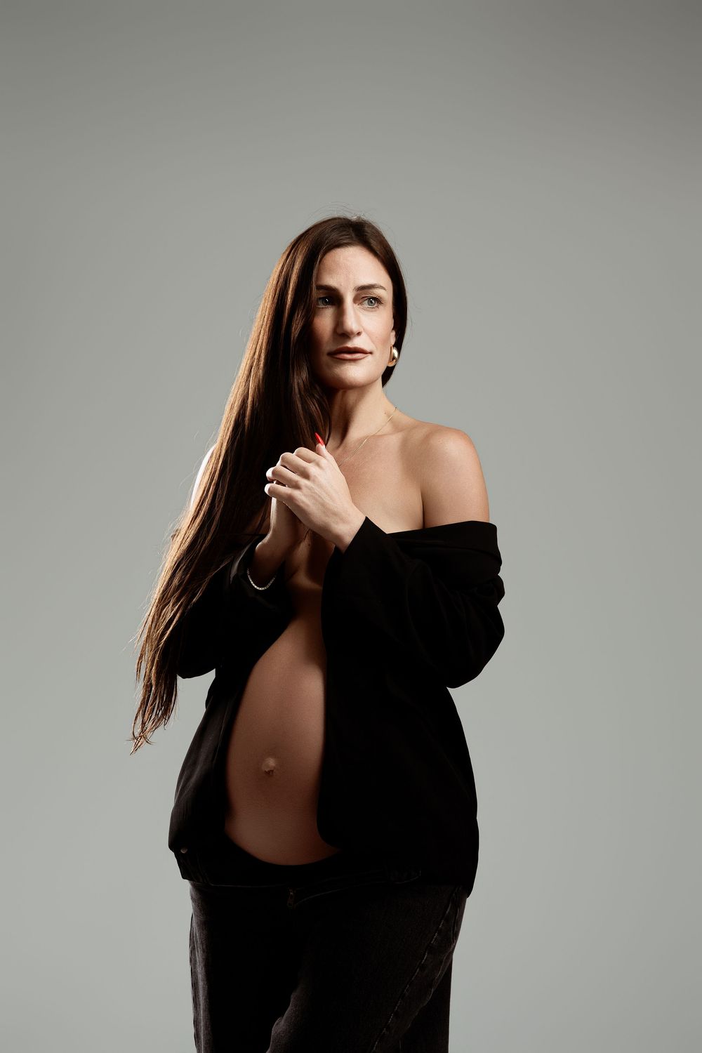 Pregnant woman in black off-shoulder jacket posing with long brown hair against gray background.
