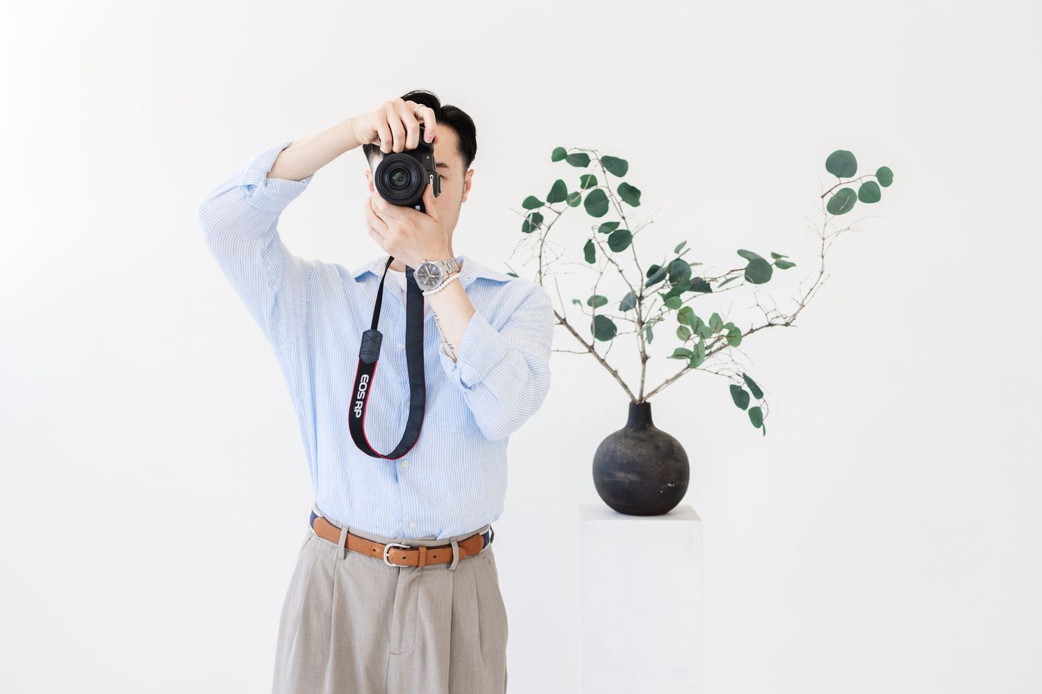 Young man in cream shirt and gray pants photographing with a camera against a light blue background.