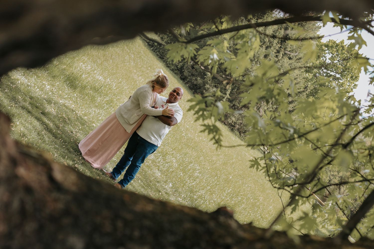An aerial view of a couple embracing on green grass, framed by tree branches overhead.