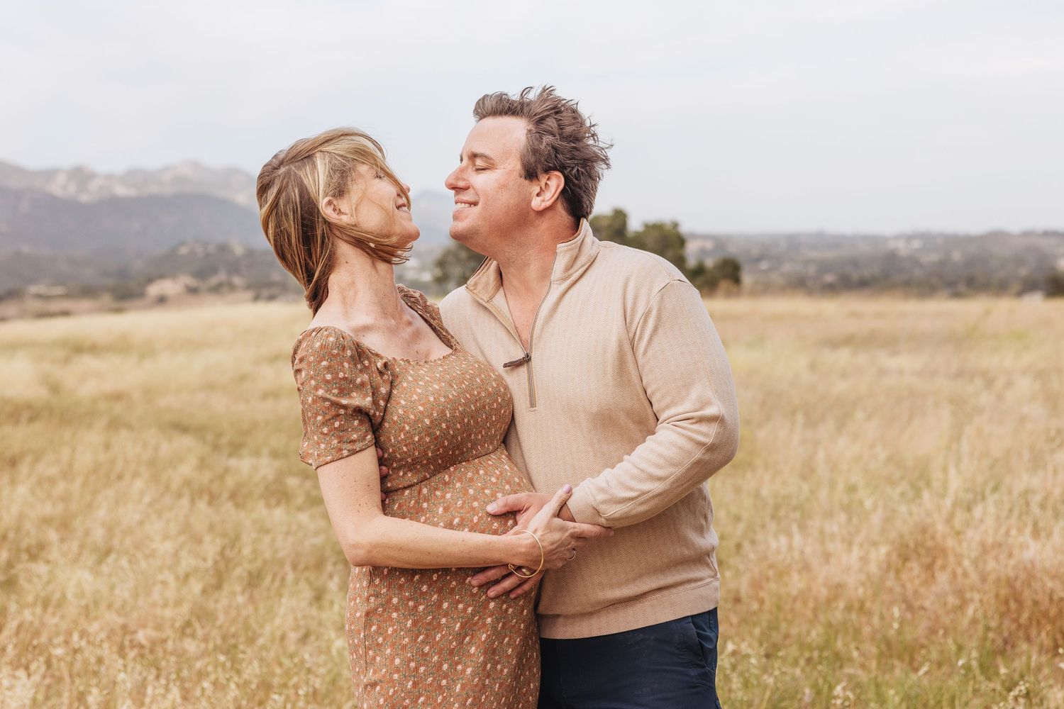 Pregnant couple embracing in field with mountains, woman in dress, man in beige sweater, sharing tender moment with maternity photographer