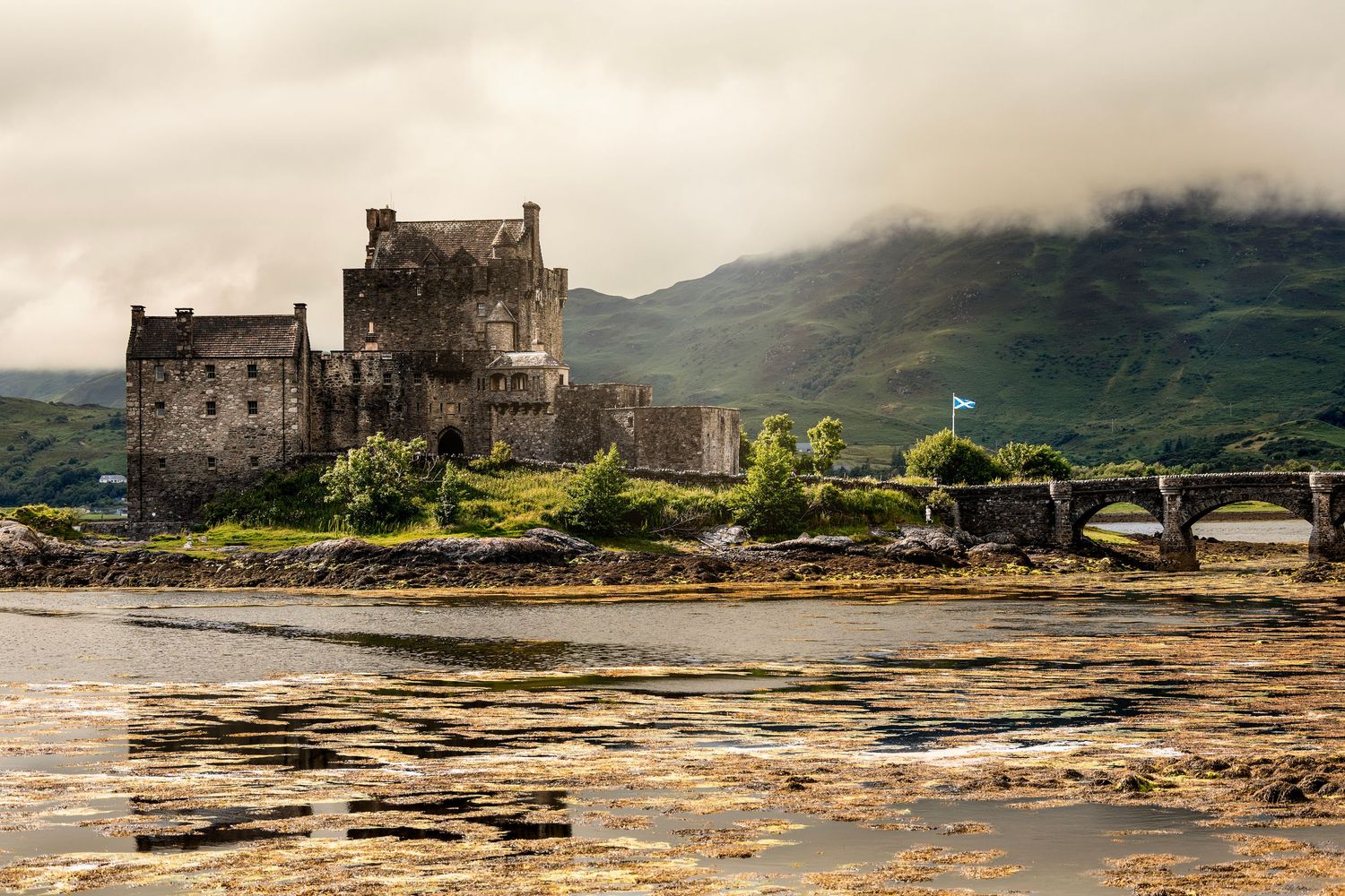 Eilean Donan Castle sits on a misty loch in the Scottish Highlands with a stone bridge and green hills behind.