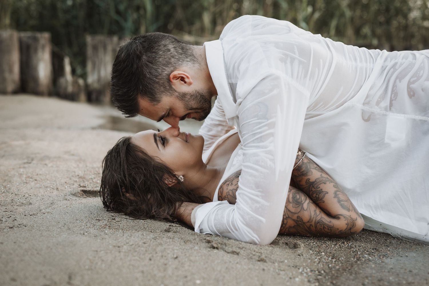 Couple de mariés allongés sur le sable mouillé, nez contre nez, lors d’une séance trash the dress