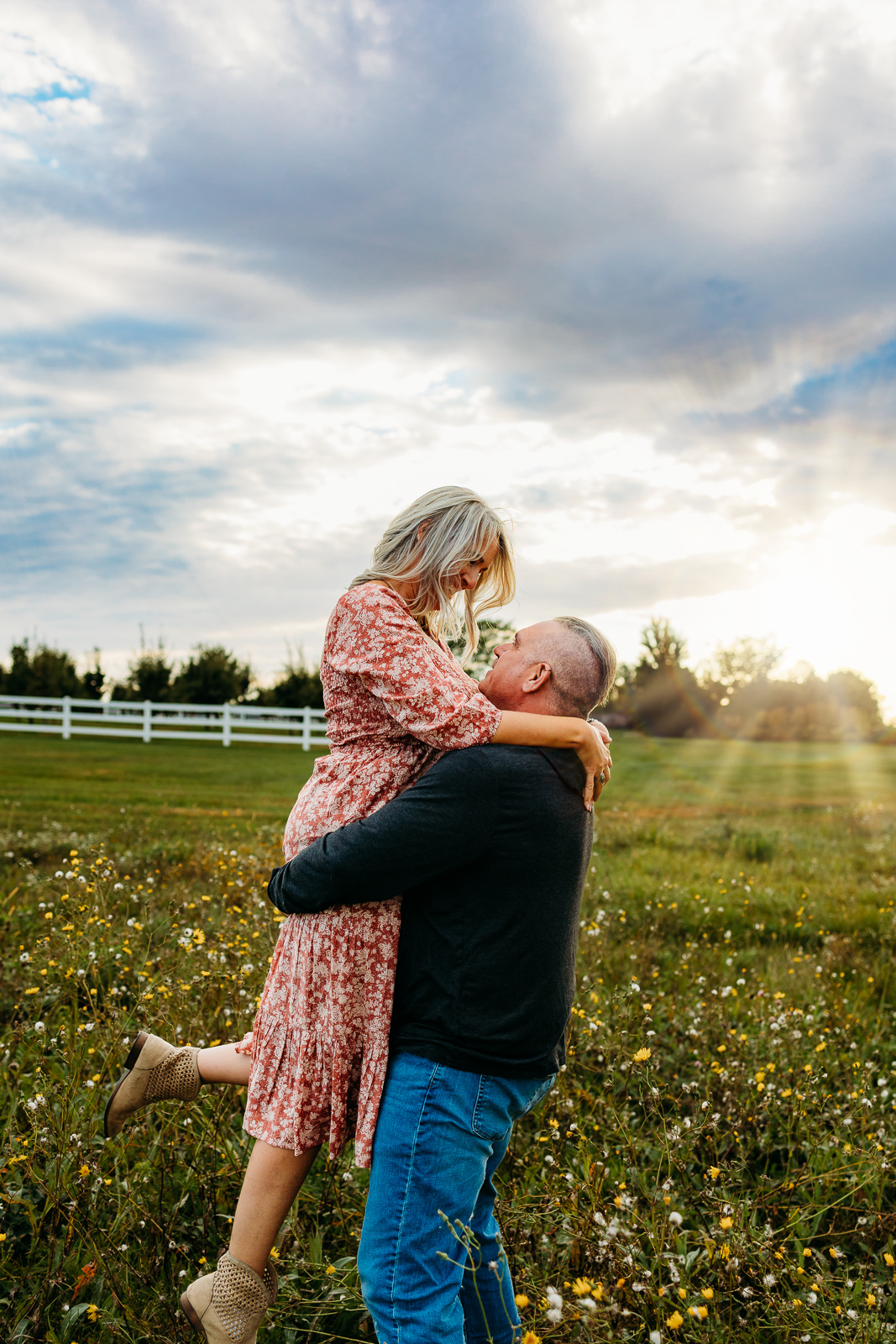 Husband lifting wife up while smiling at each other. The sun is shining in the background.