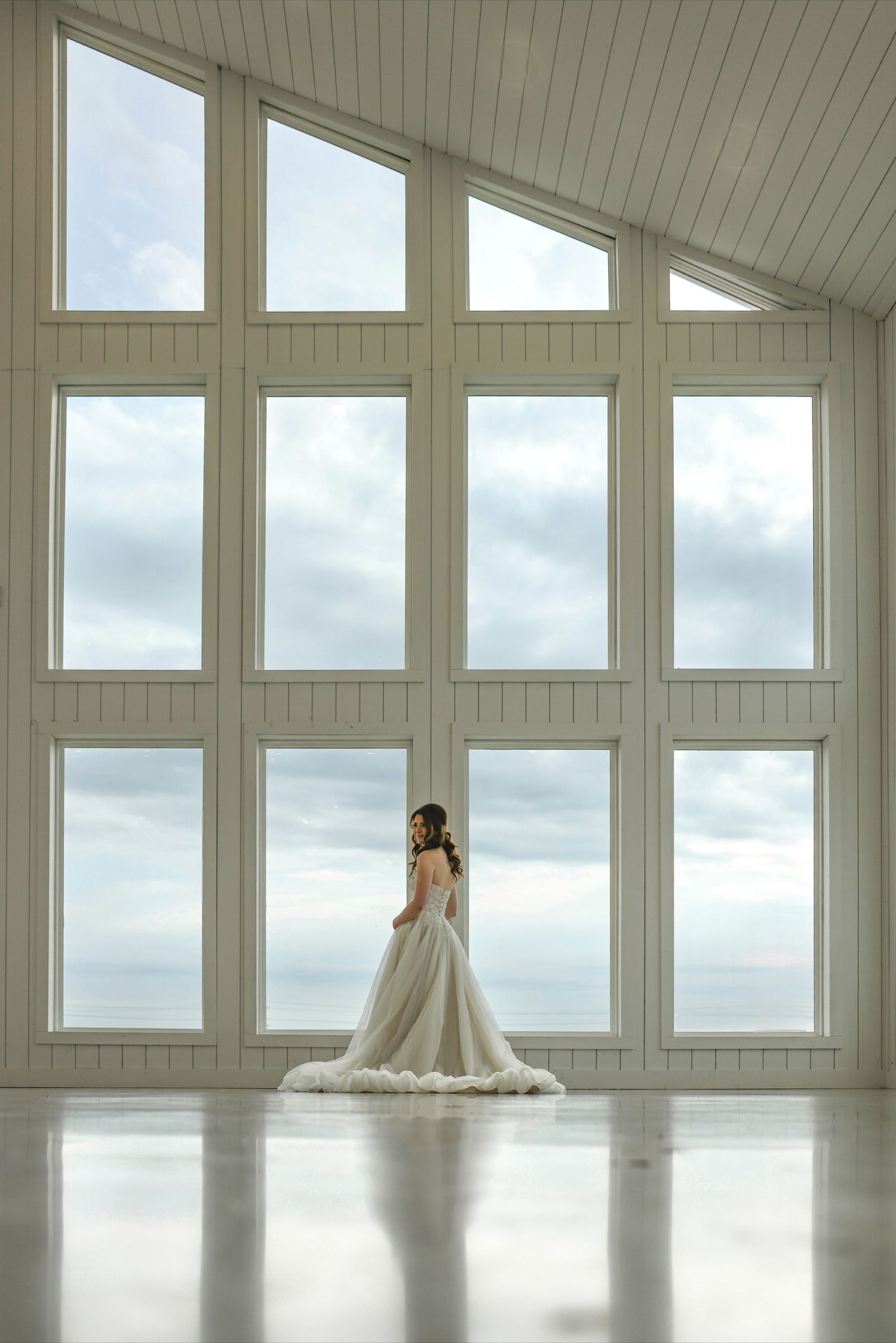 Bride in a flowing white ball gown stands before large floor-to-ceiling windows overlooking a cloudy sky in an elegant venue.